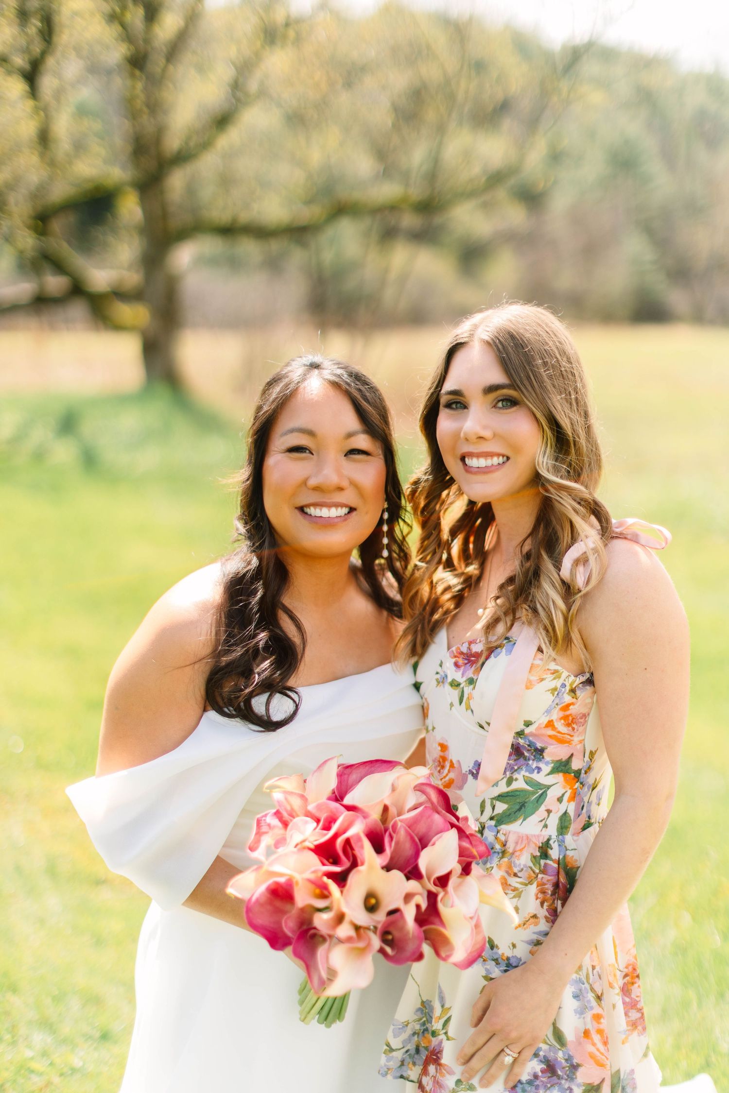 Smiling bride in white off-shoulder gown with bridesmaid in floral dress holding pink calla lily bouquet outdoors.