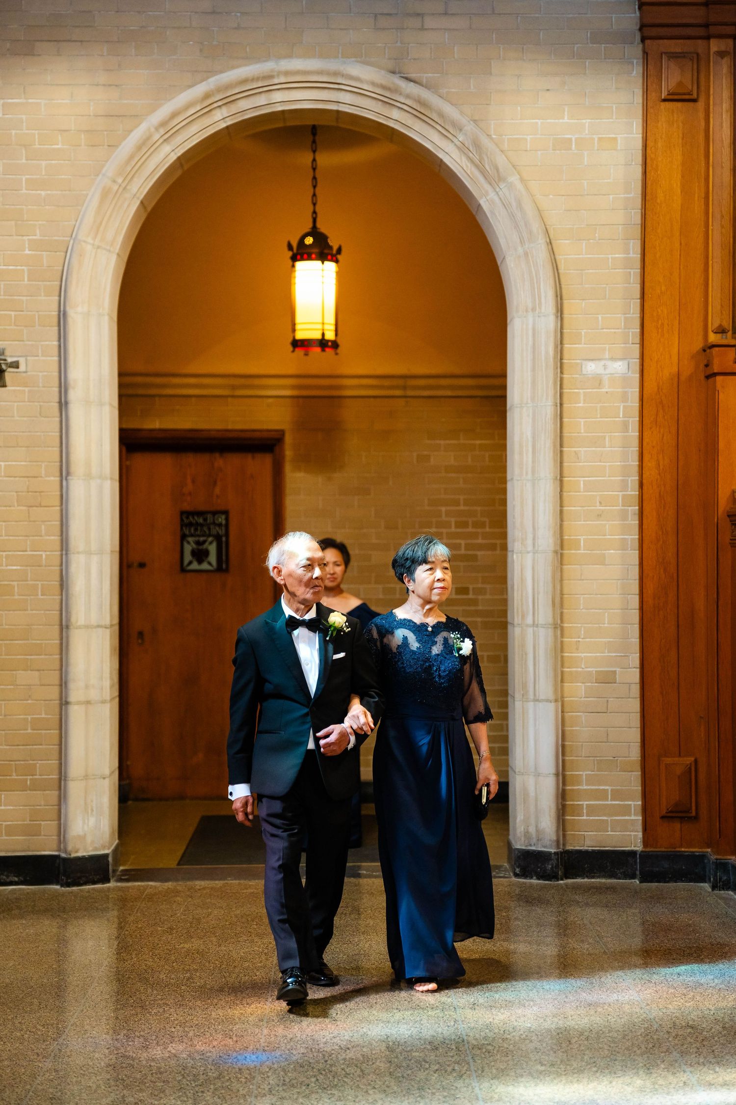 Elderly Asian couple in formal attire walking through a stone archway at a wedding ceremony.