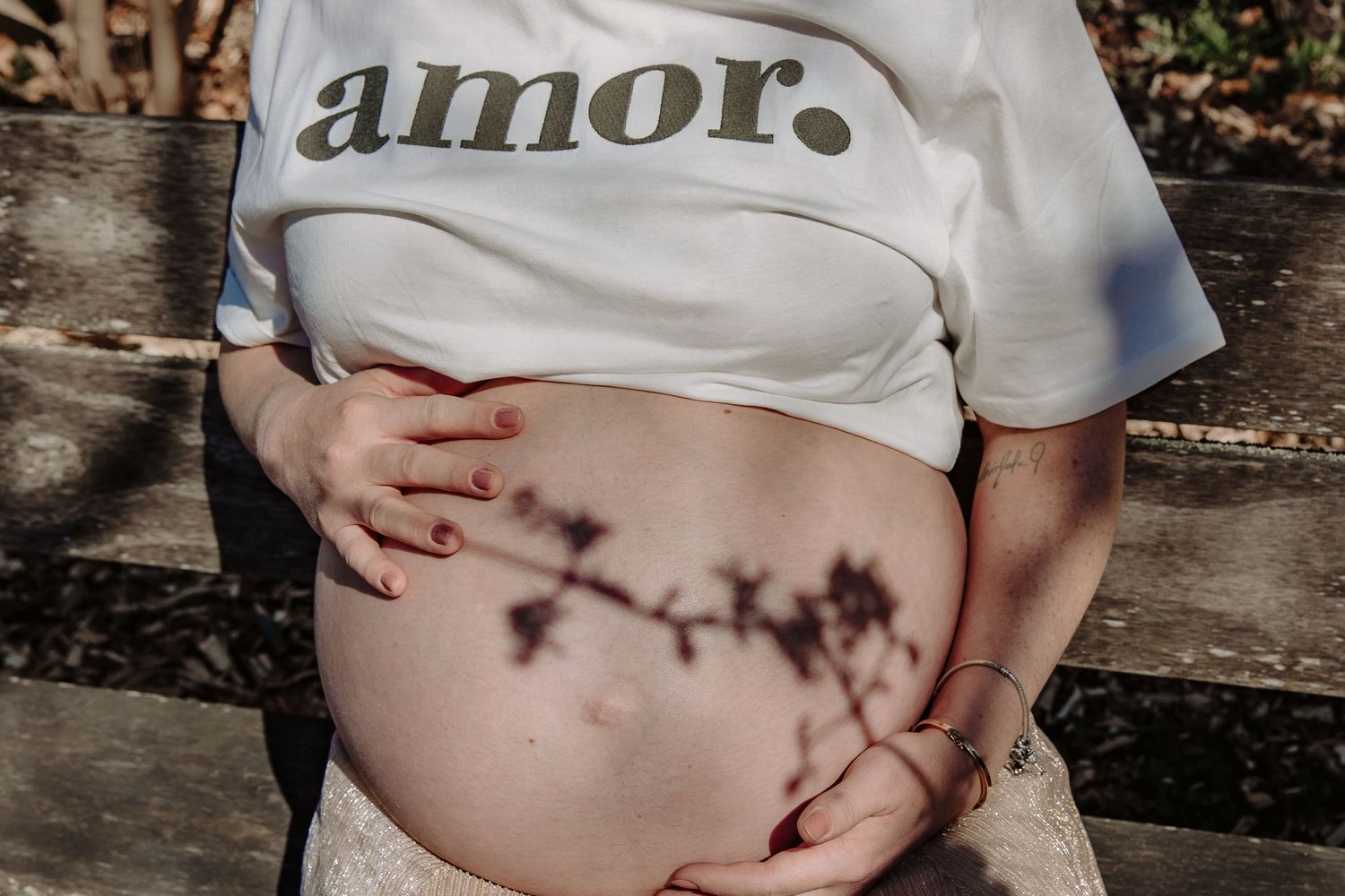 A pregnant person wearing a white t-shirt with 'amor' text sitting on a wooden bench.