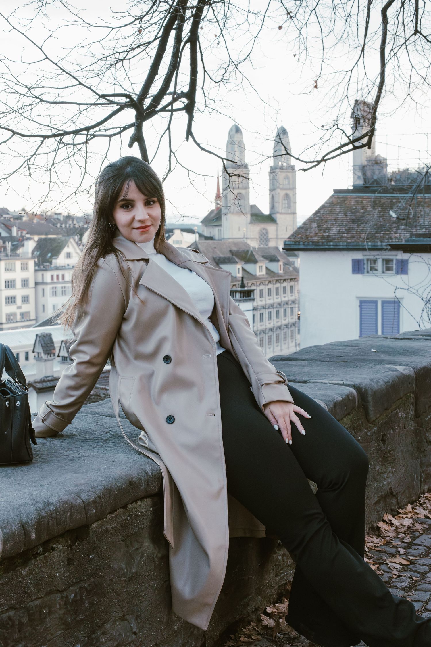 A person in a beige trench coat poses along a stone wall overlooking Zurich's historic skyline.