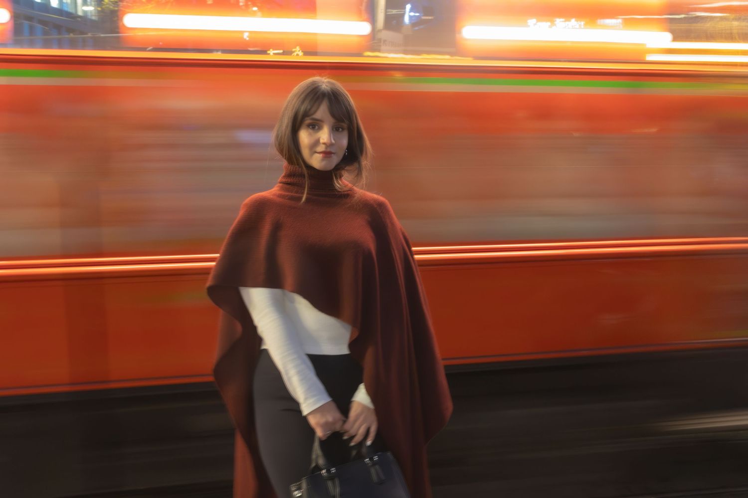 A figure in a burgundy poncho stands still as red transit vehicles blur past in the background.