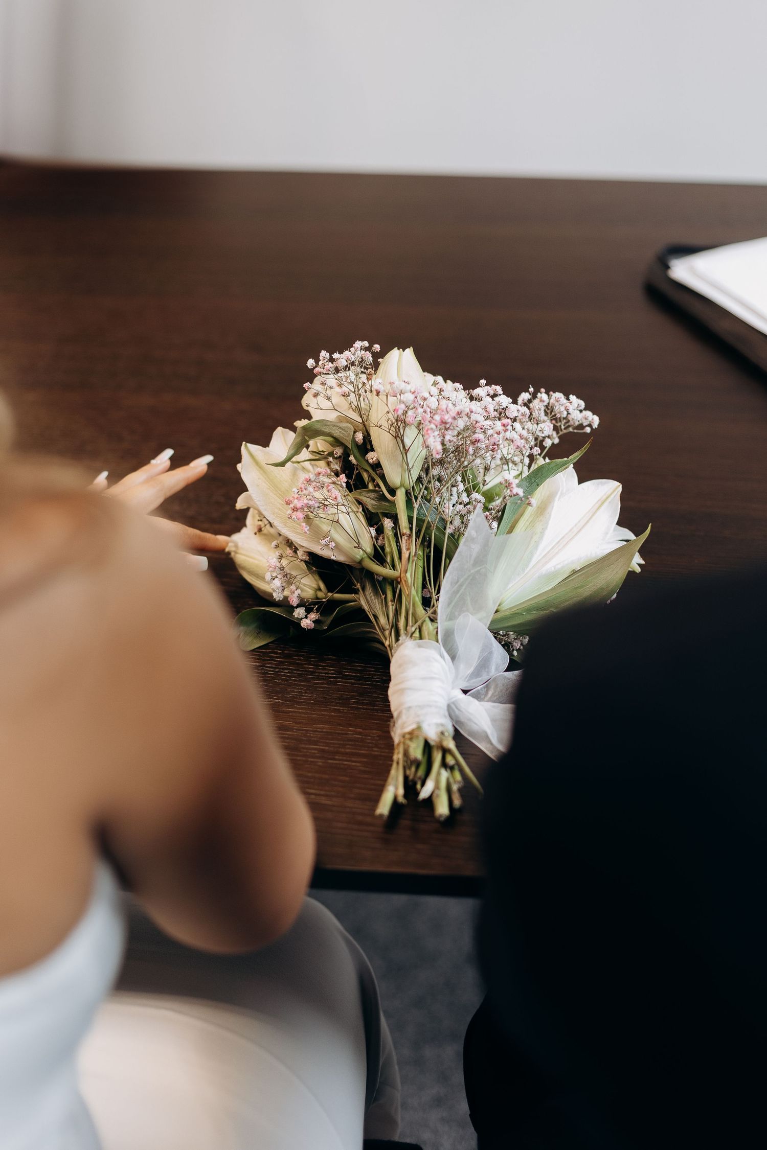 A delicate wedding bouquet with white lilies and pink flowers tied with a white ribbon sits on a wooden table.
