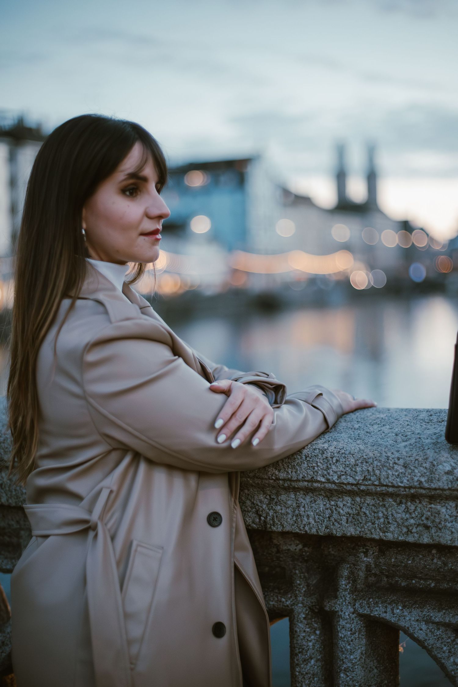 Person in beige trench coat leaning on stone railing with city view at sunset.
