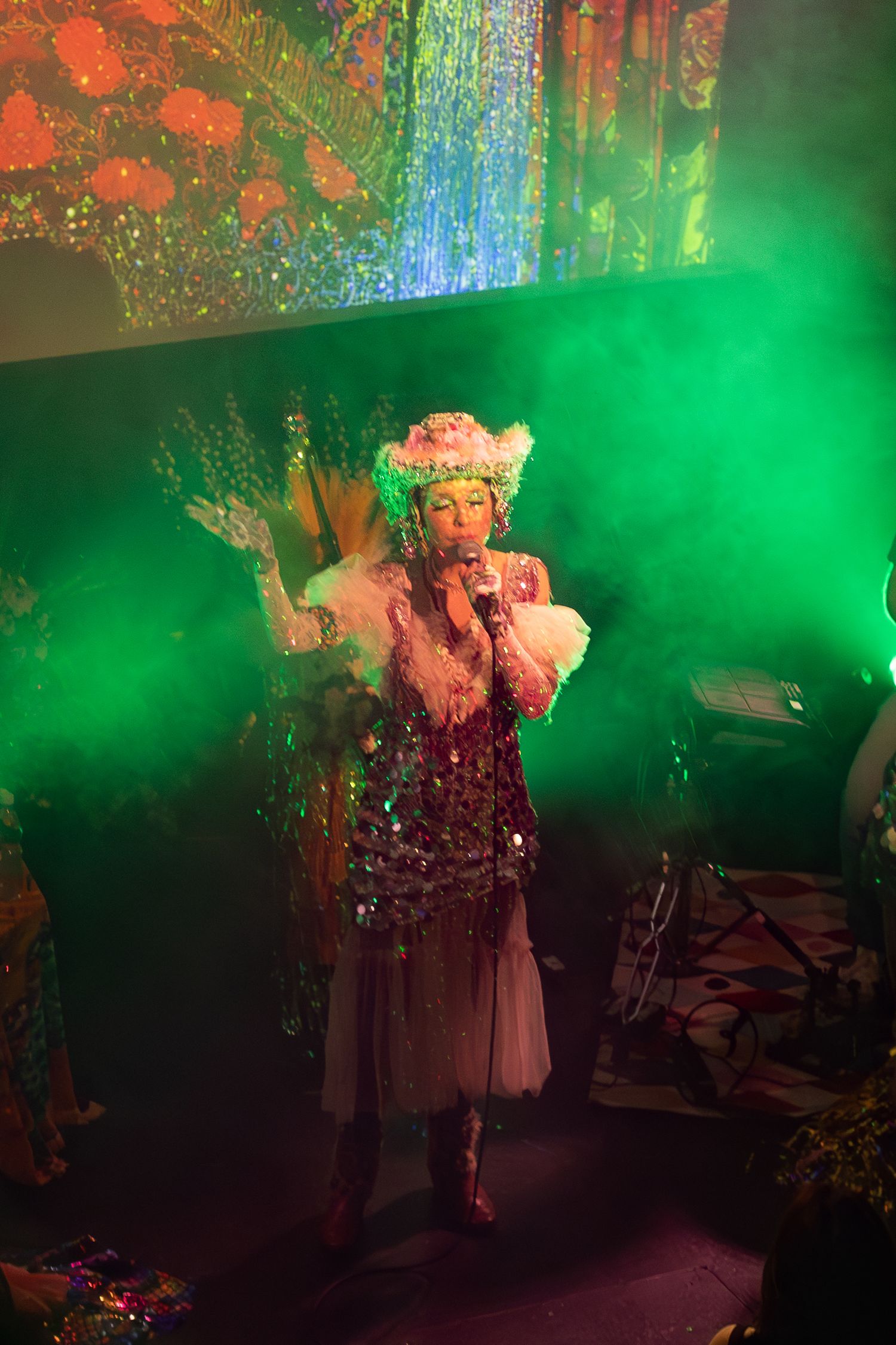 A performer in vintage-inspired costume performs under green stage lighting with colorful projections.