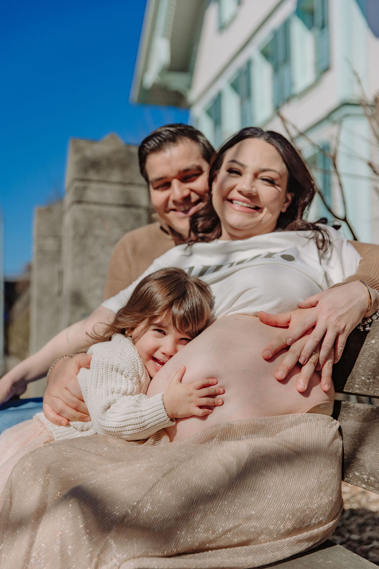 A family embraces while sitting outdoors, with a child hugging a pregnant belly.