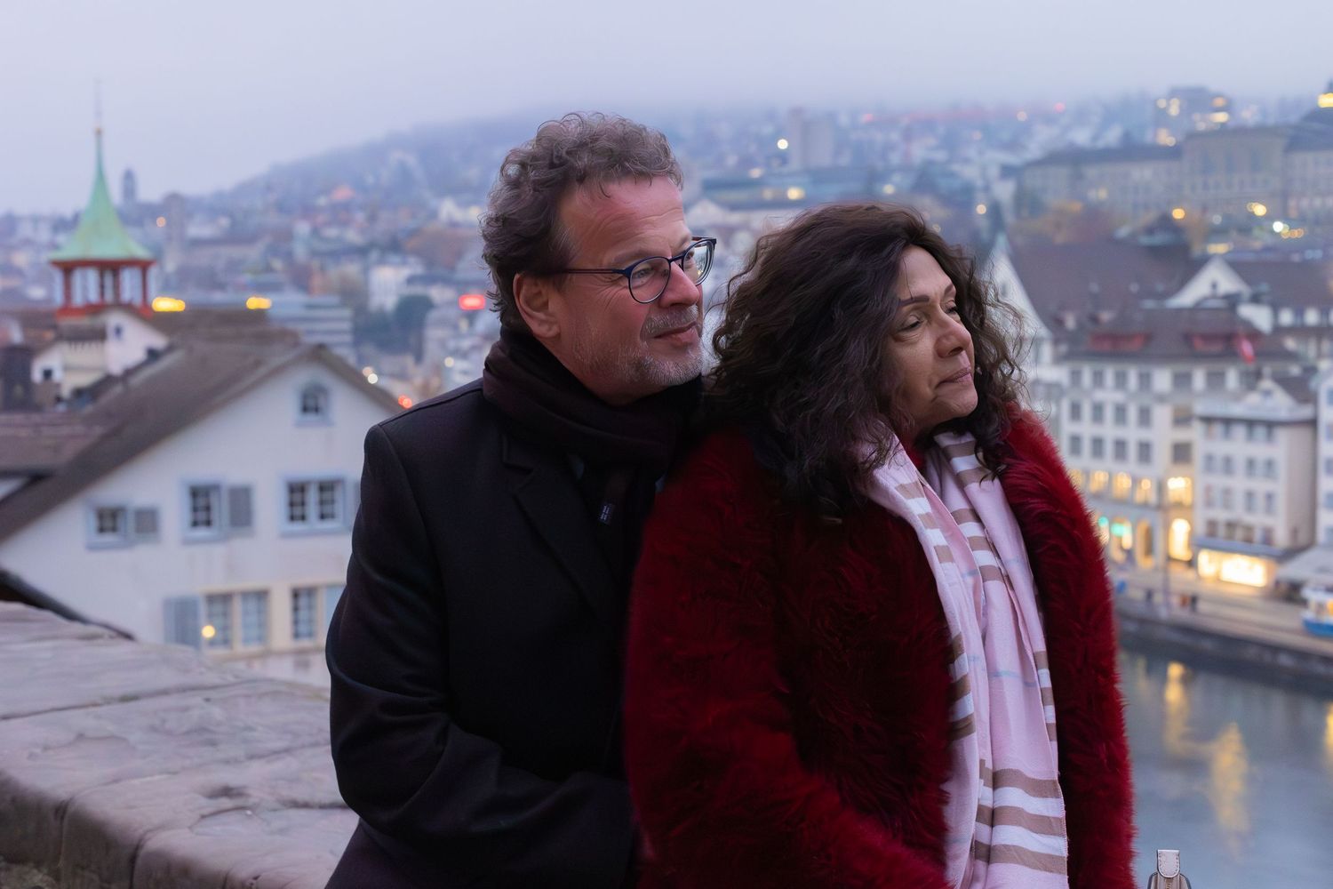 A couple stands together at dusk overlooking the Zurich cityscape and river.