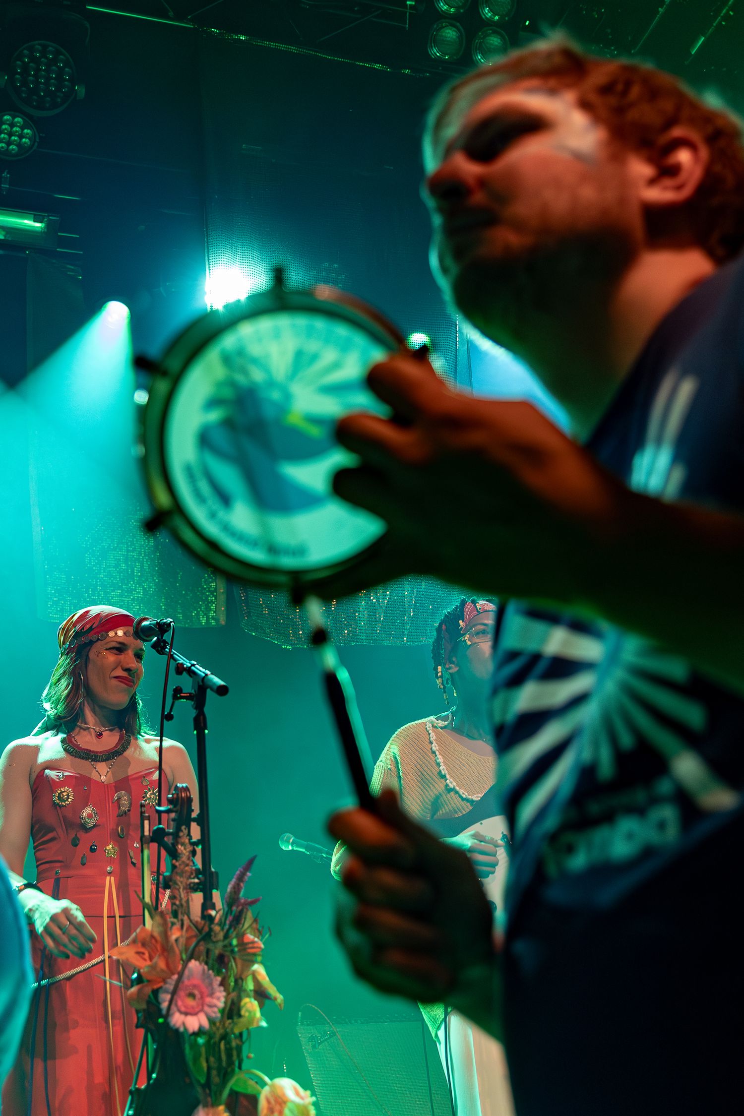A drum performer plays on stage bathed in turquoise lighting while another musician sings at a concert.