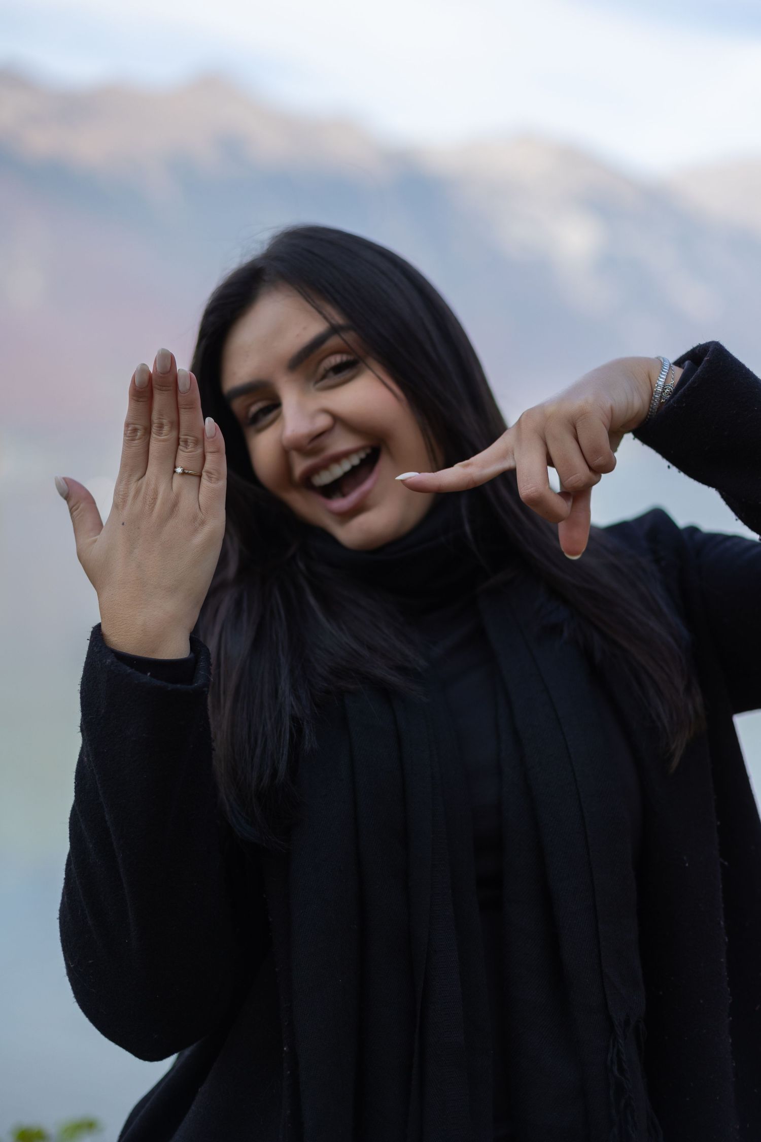A person in black clothing makes playful hand gestures against a mountain backdrop.