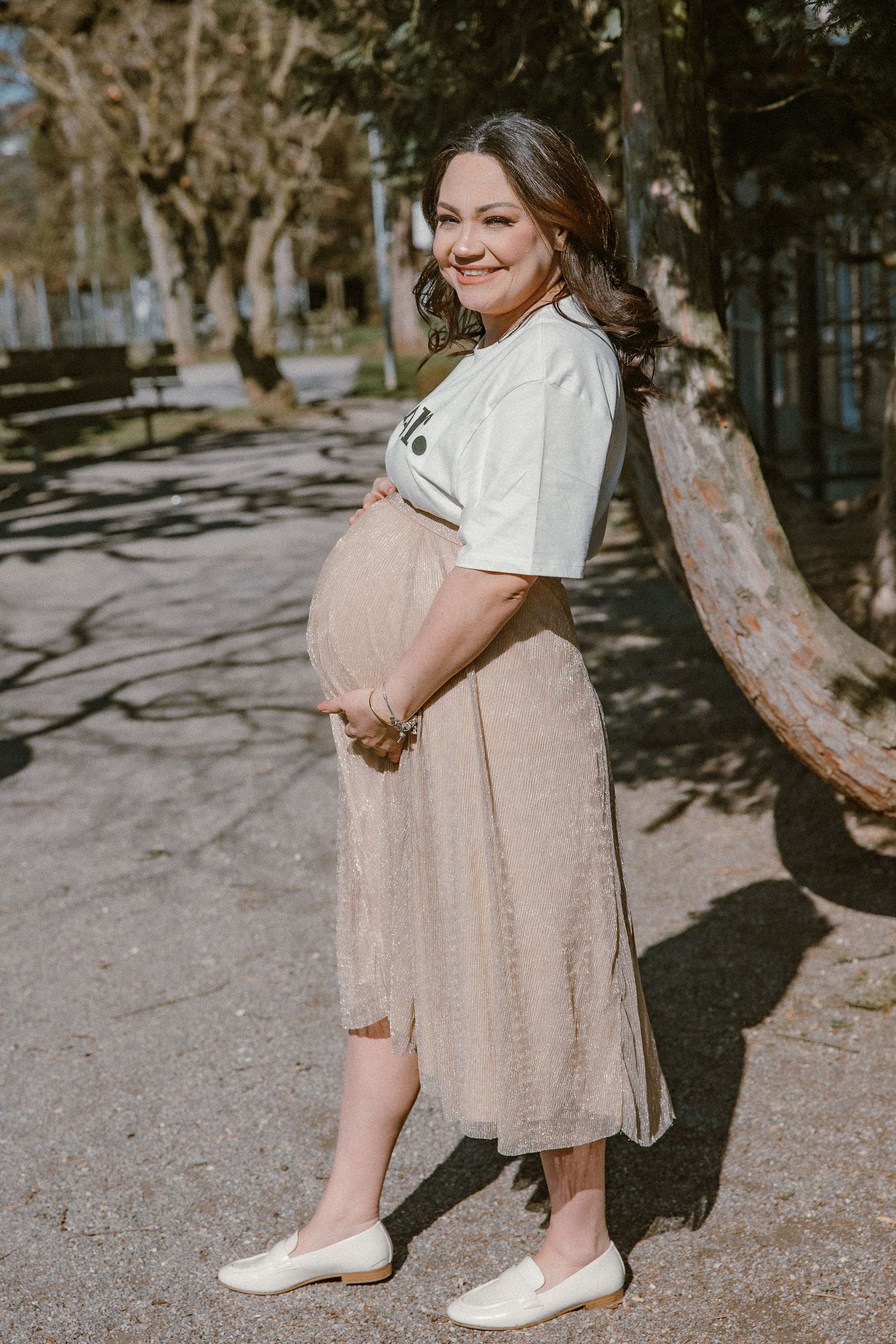 Pregnant woman in white t-shirt and beige skirt poses confidently on tree-lined path.