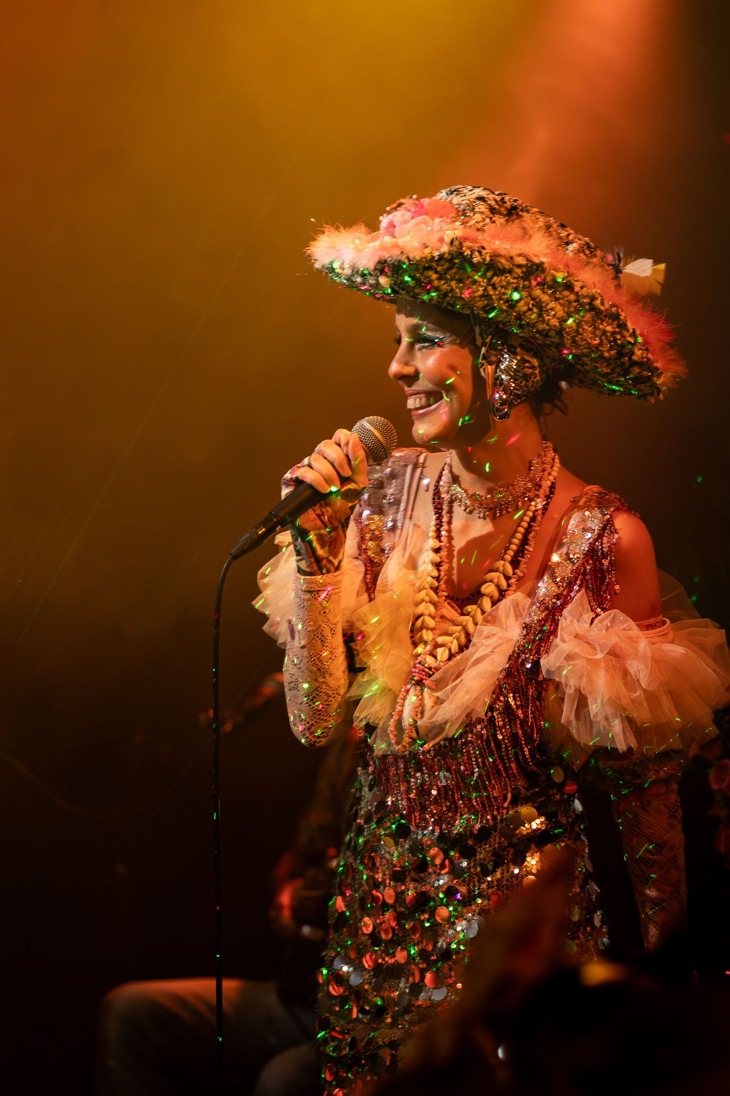A performer in an elaborate feathered costume and headpiece sings on stage under warm lighting.