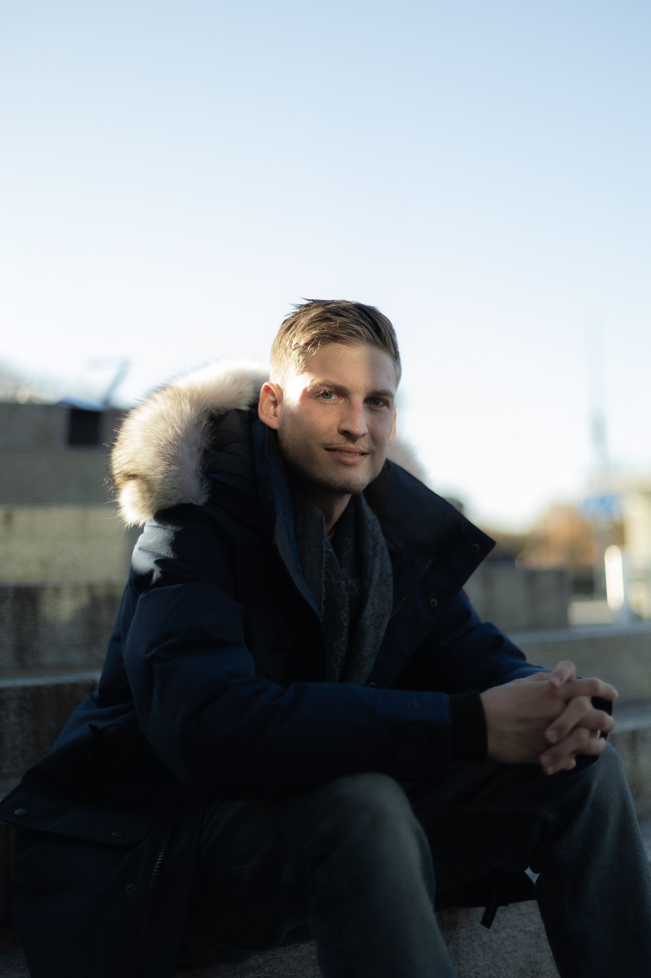 Figure in winter coat with fur hood sitting outdoors against bright sky.