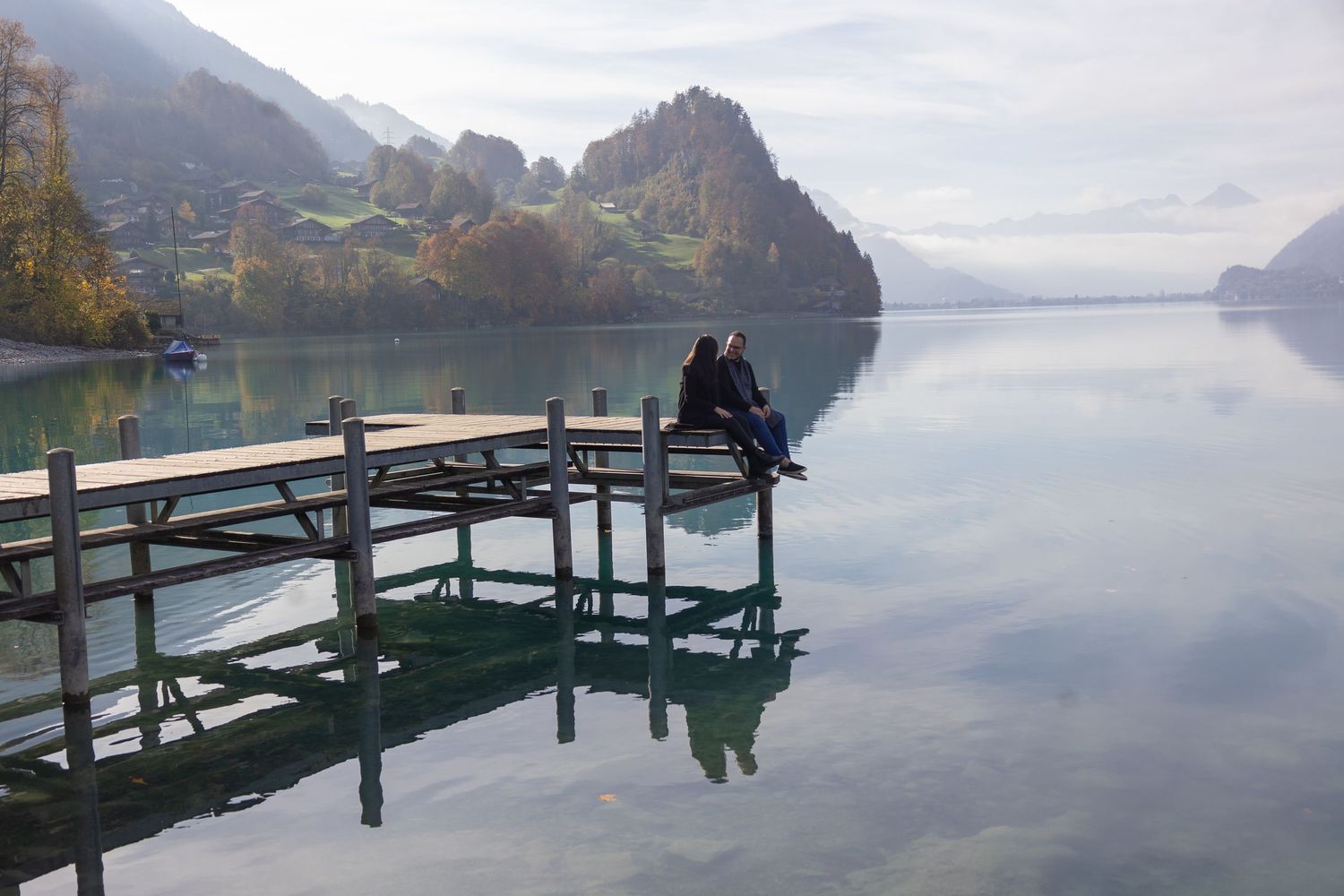 Two people sit close together on wooden dock overlooking tranquil mountain lake.