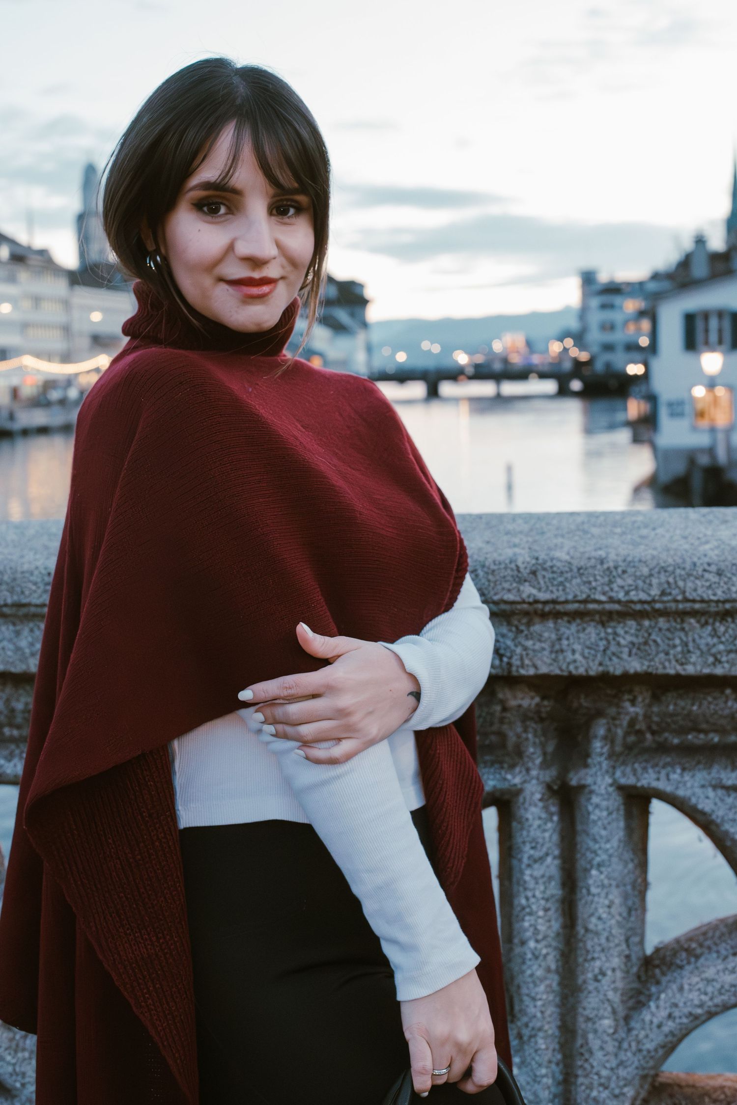 A person in a burgundy wrap poses against a waterfront cityscape at twilight.