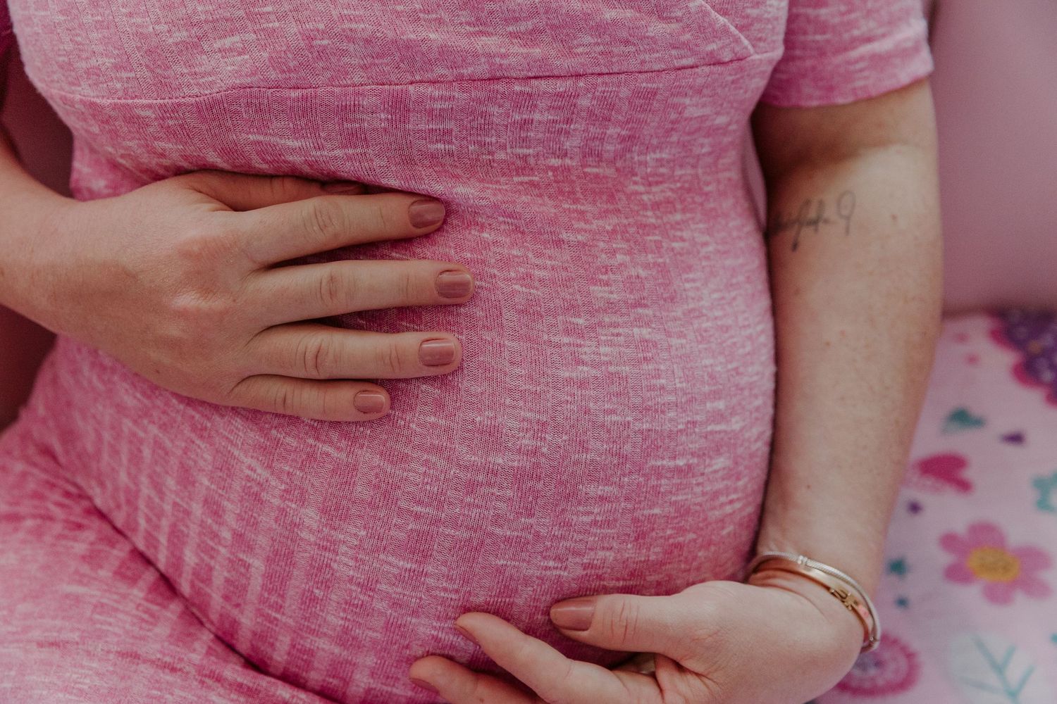 Close-up of hands gently resting on a pregnant belly in a pink dress.