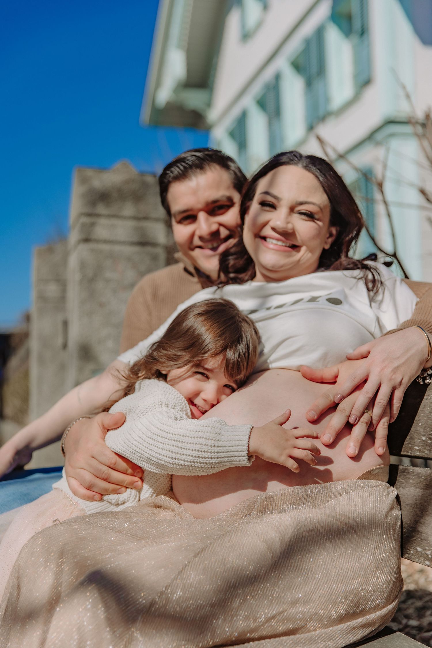 A family shares a joyful embrace outdoors on a sunny day with a stone building in the background.