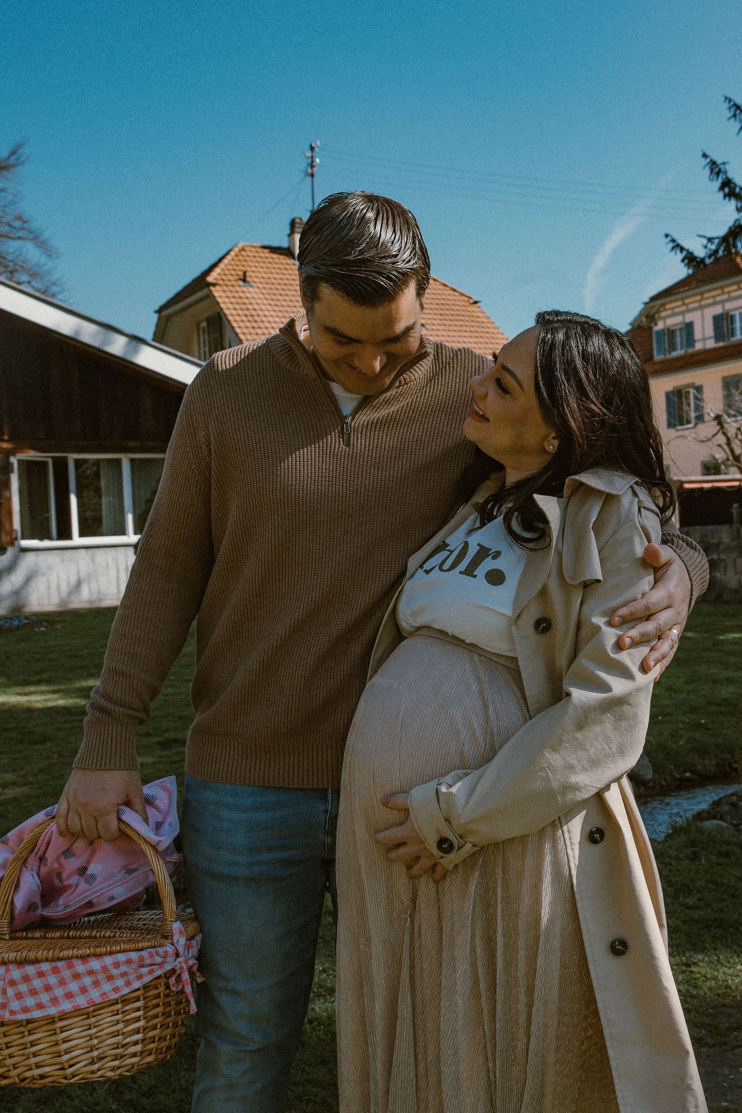 A couple stands together outdoors with a picnic basket, surrounded by houses on a sunny day.