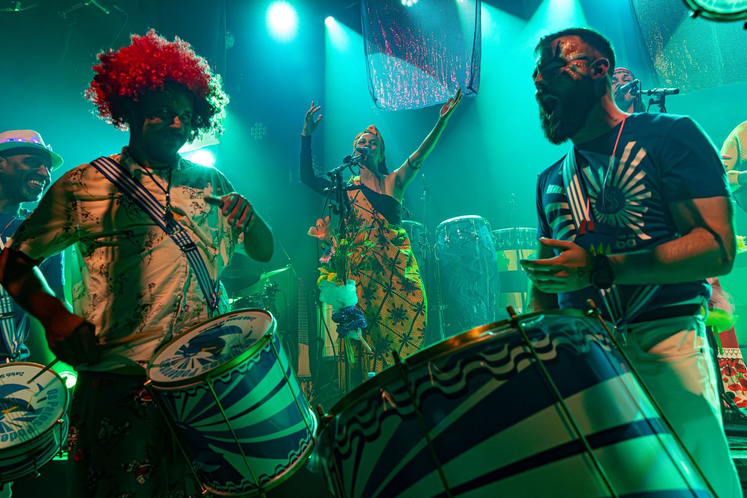 Drummers with festive costumes and red hair accessories perform energetically under teal stage lights.