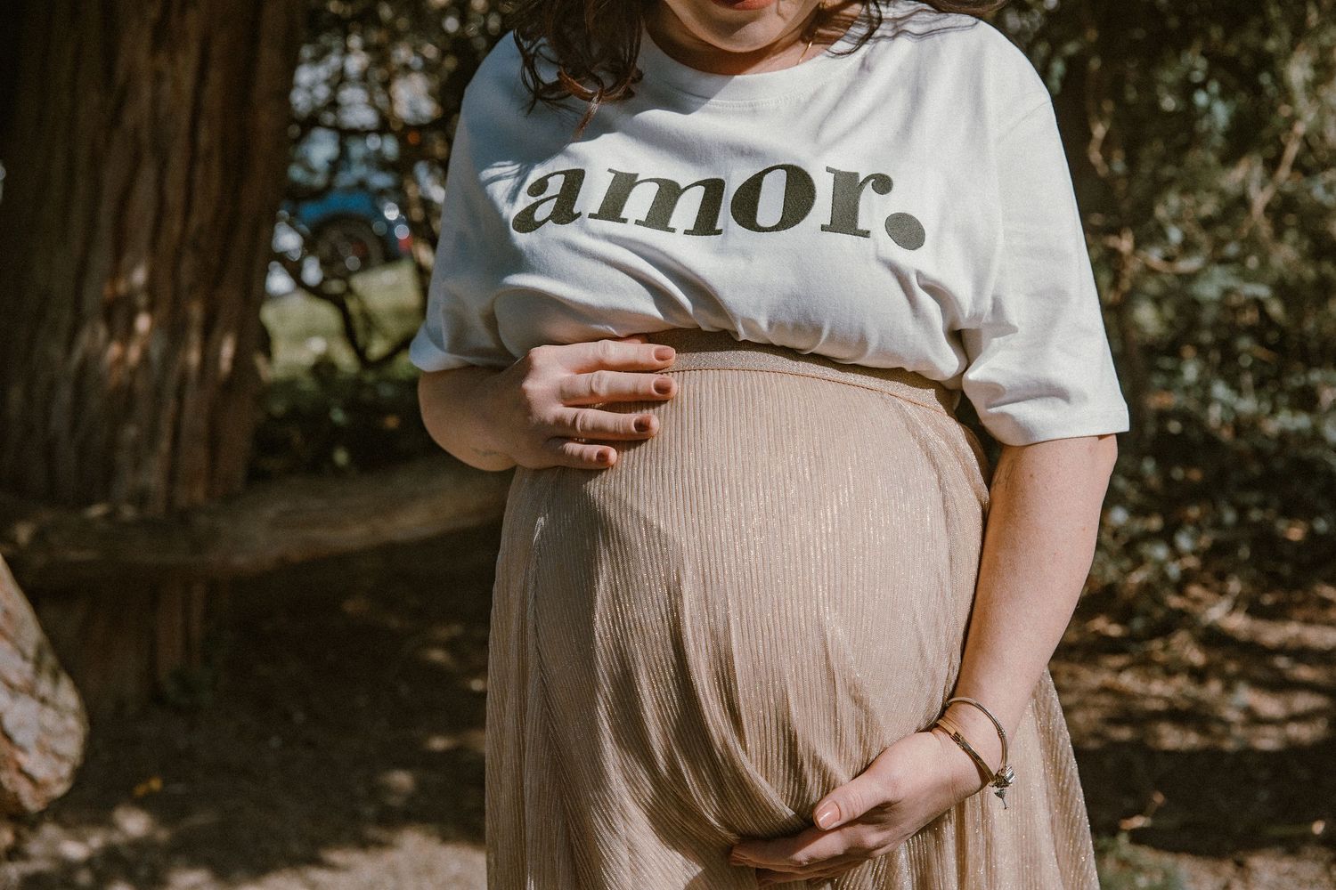 Pregnant woman wearing 'amor' t-shirt gently holds her baby bump in outdoor setting.