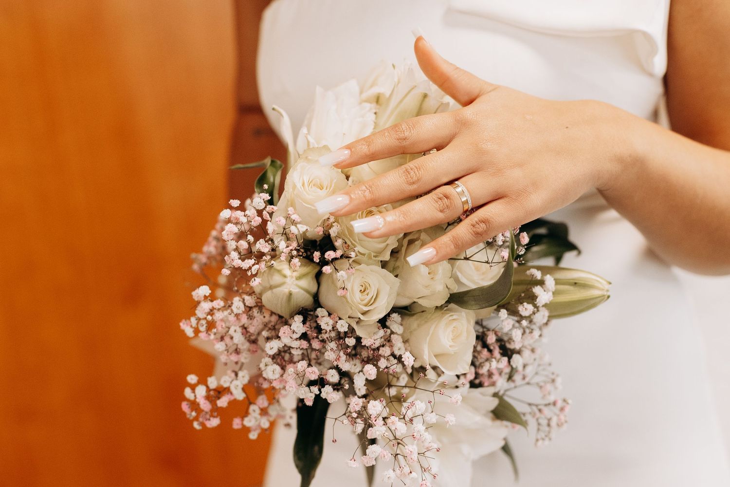 A bridal bouquet of white roses and baby's breath held against a white dress with a warm orange background.
