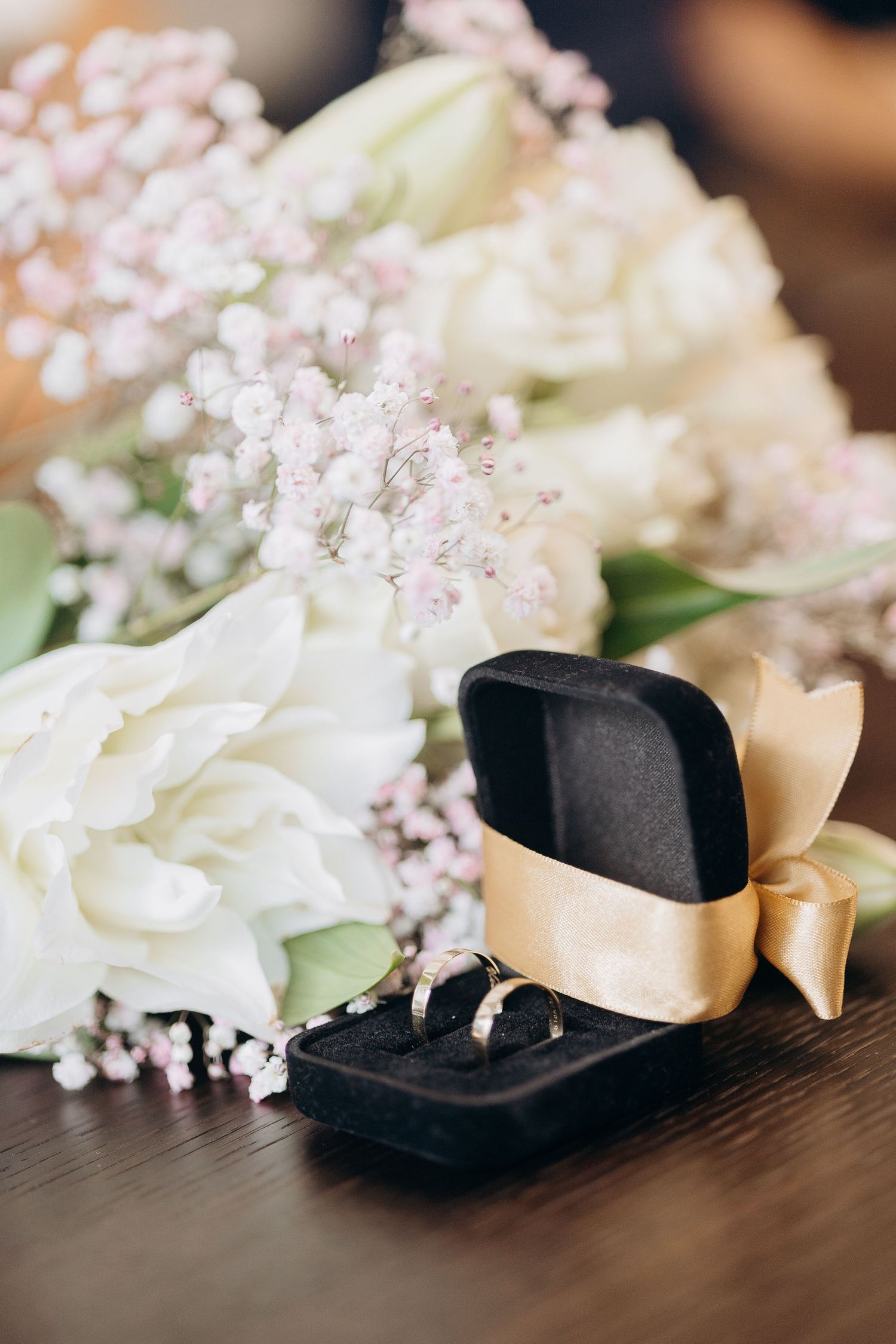 Wedding rings in black box with ribbon beside white and pink floral arrangement.