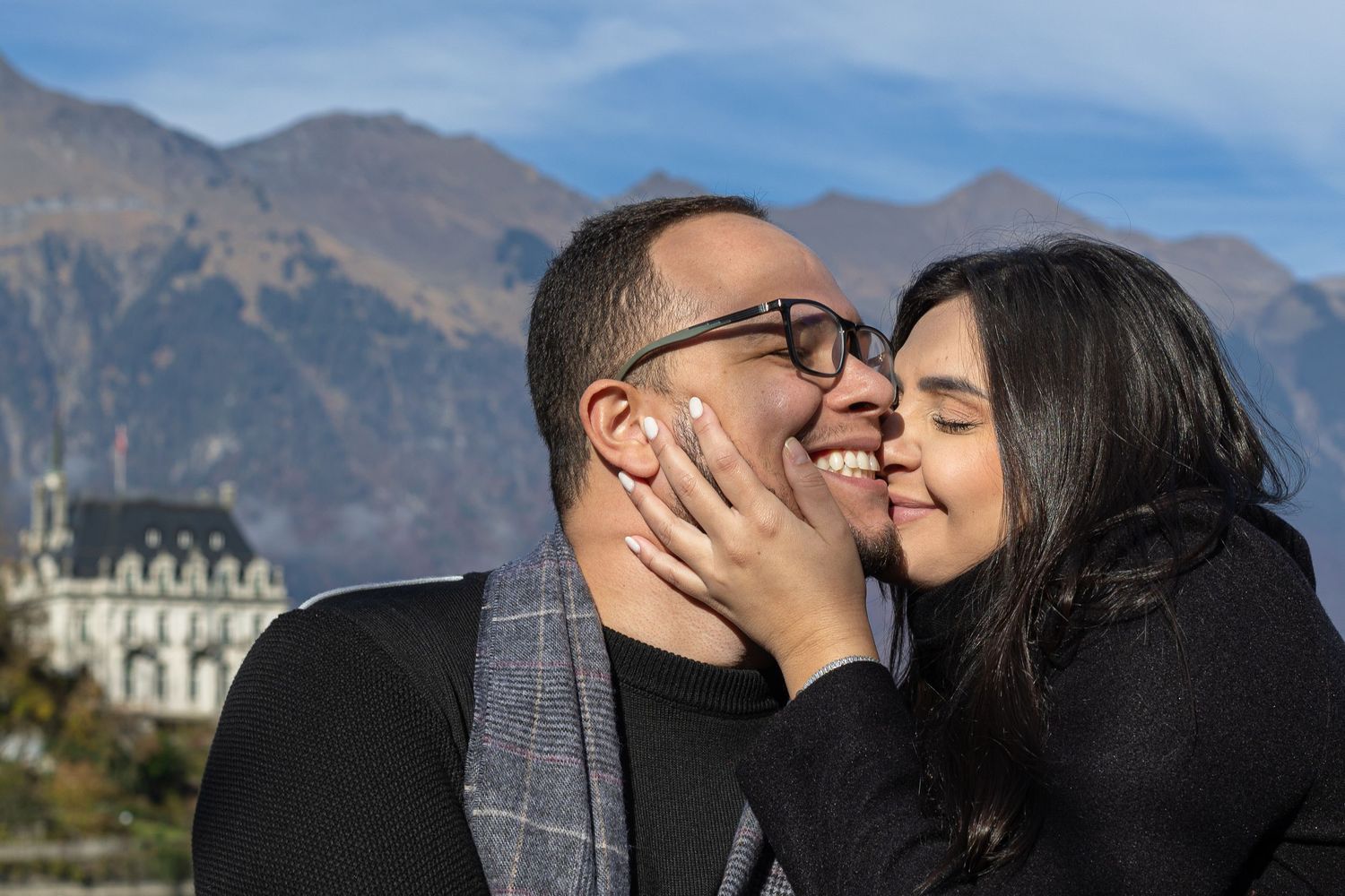 A romantic couple shares an intimate moment with a scenic mountain lake and chateau backdrop.