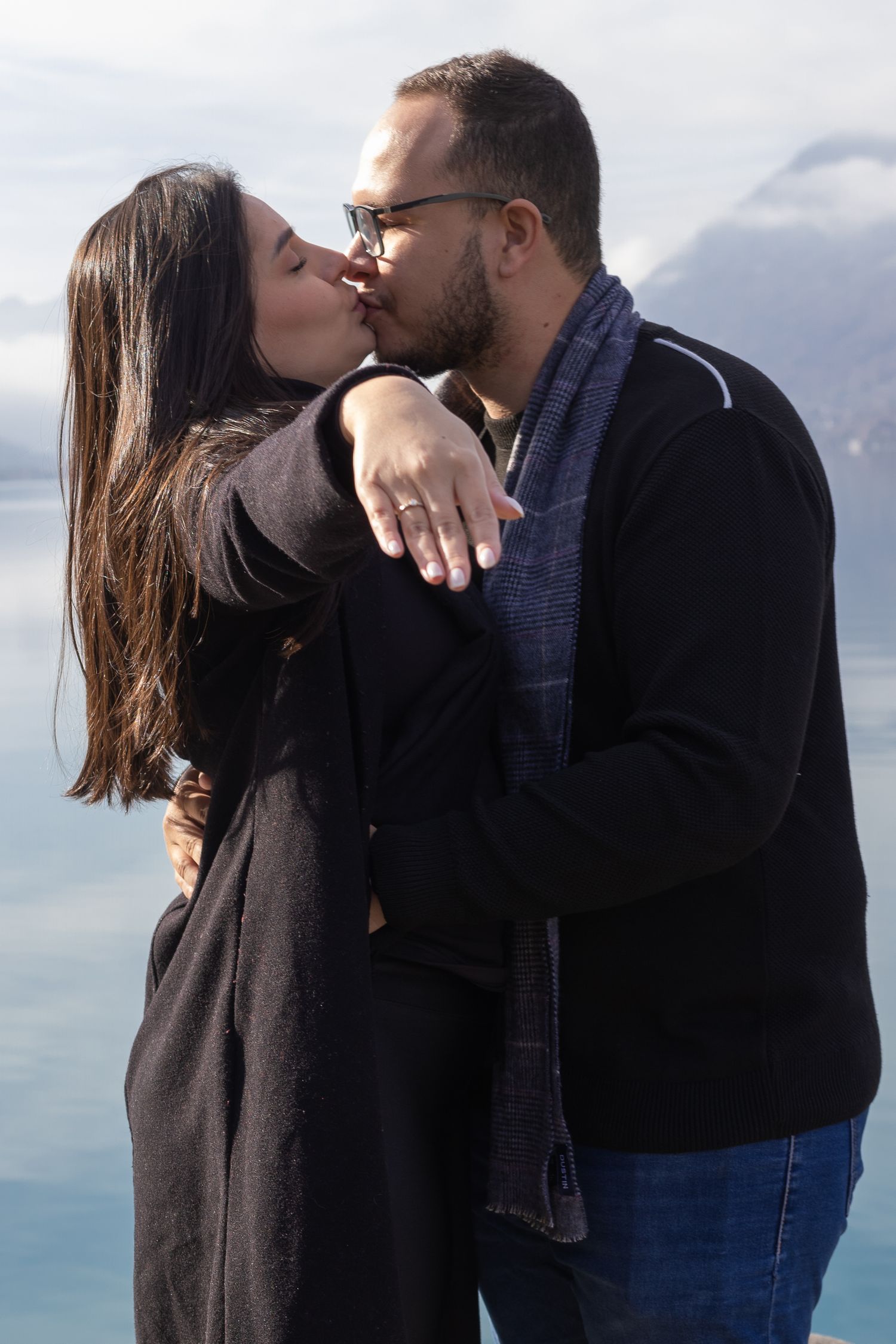 Romantic moment shared between couple on dock with ring displayed against mountain backdrop.