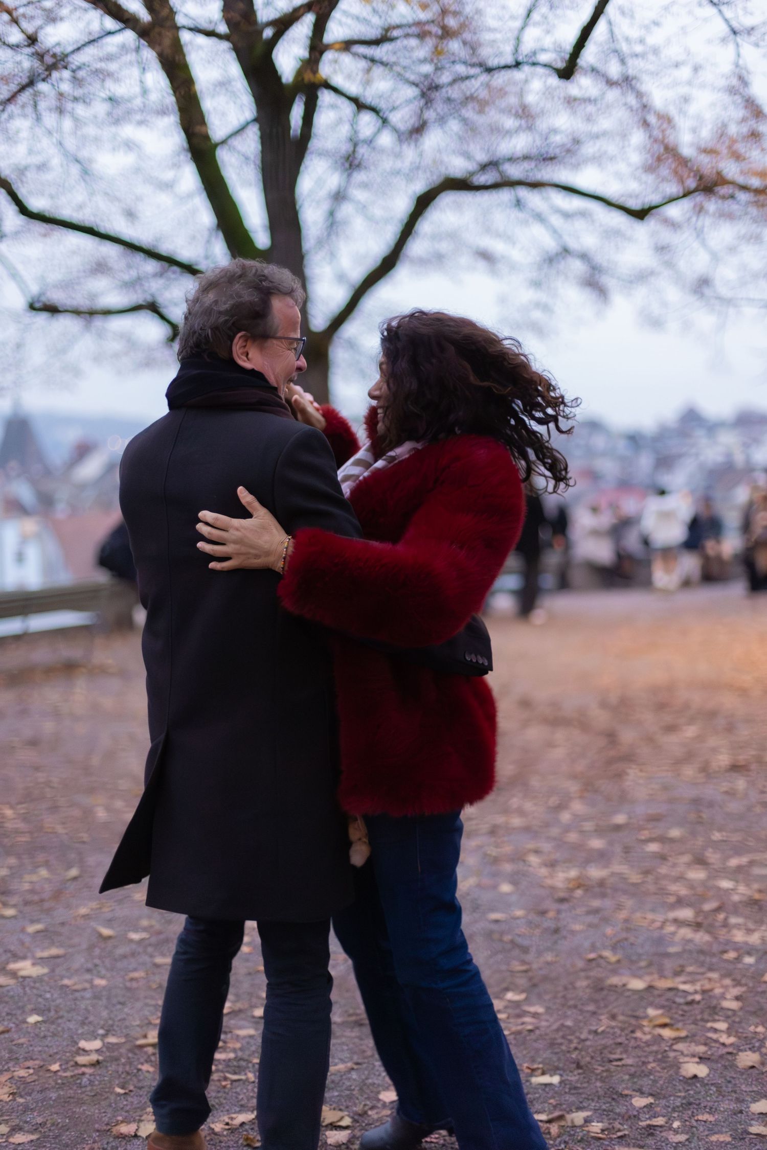 Two people embrace on an autumn path with bare trees and city views.