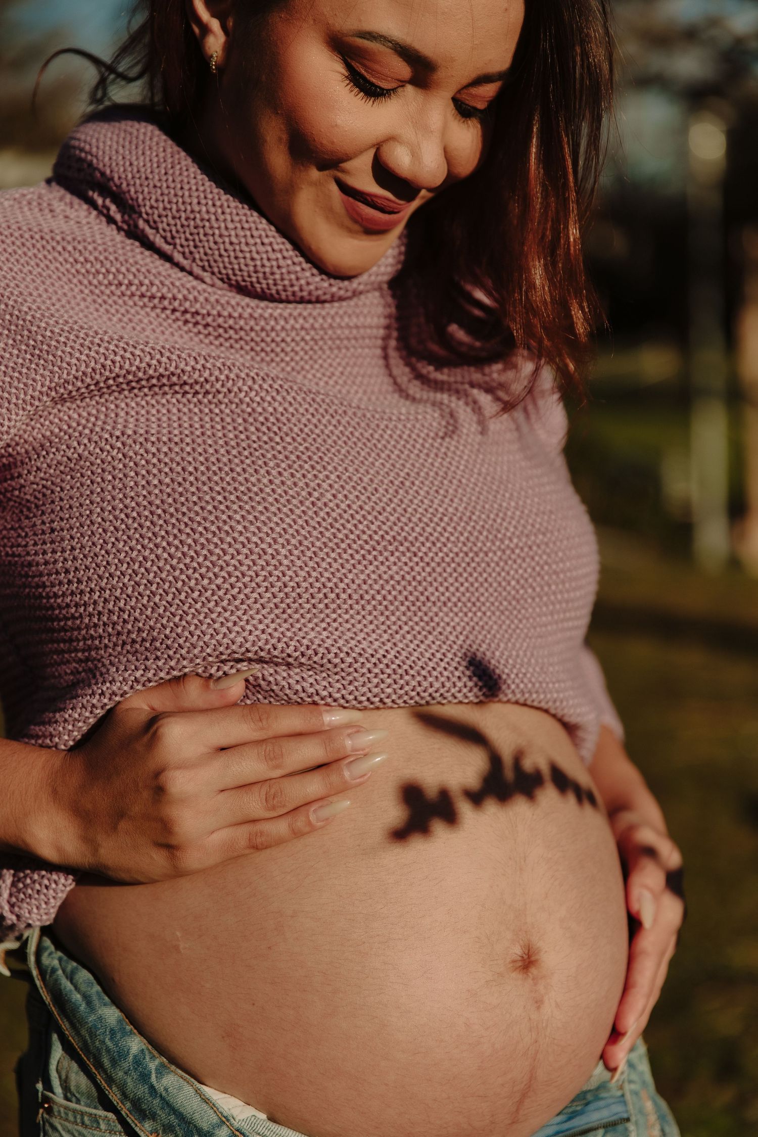 Maternity photo showing pregnant belly with writing while wearing pink knit sweater outdoors.