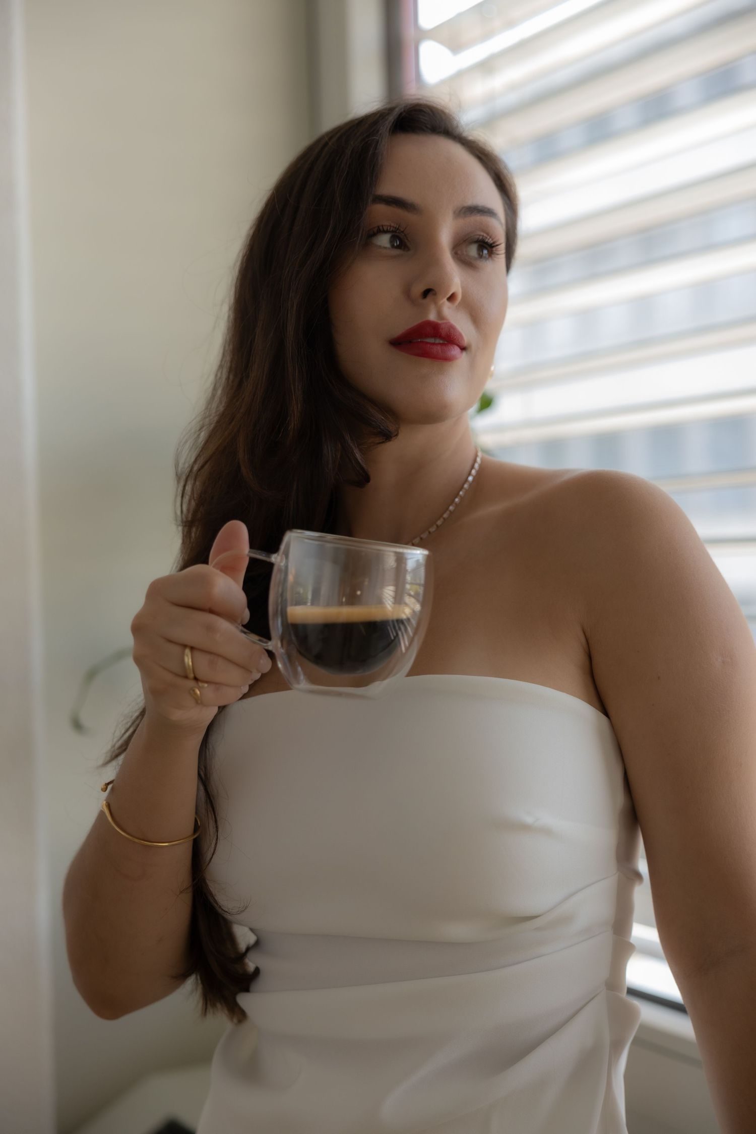Person drinking from glass cup by window in elegant white attire.