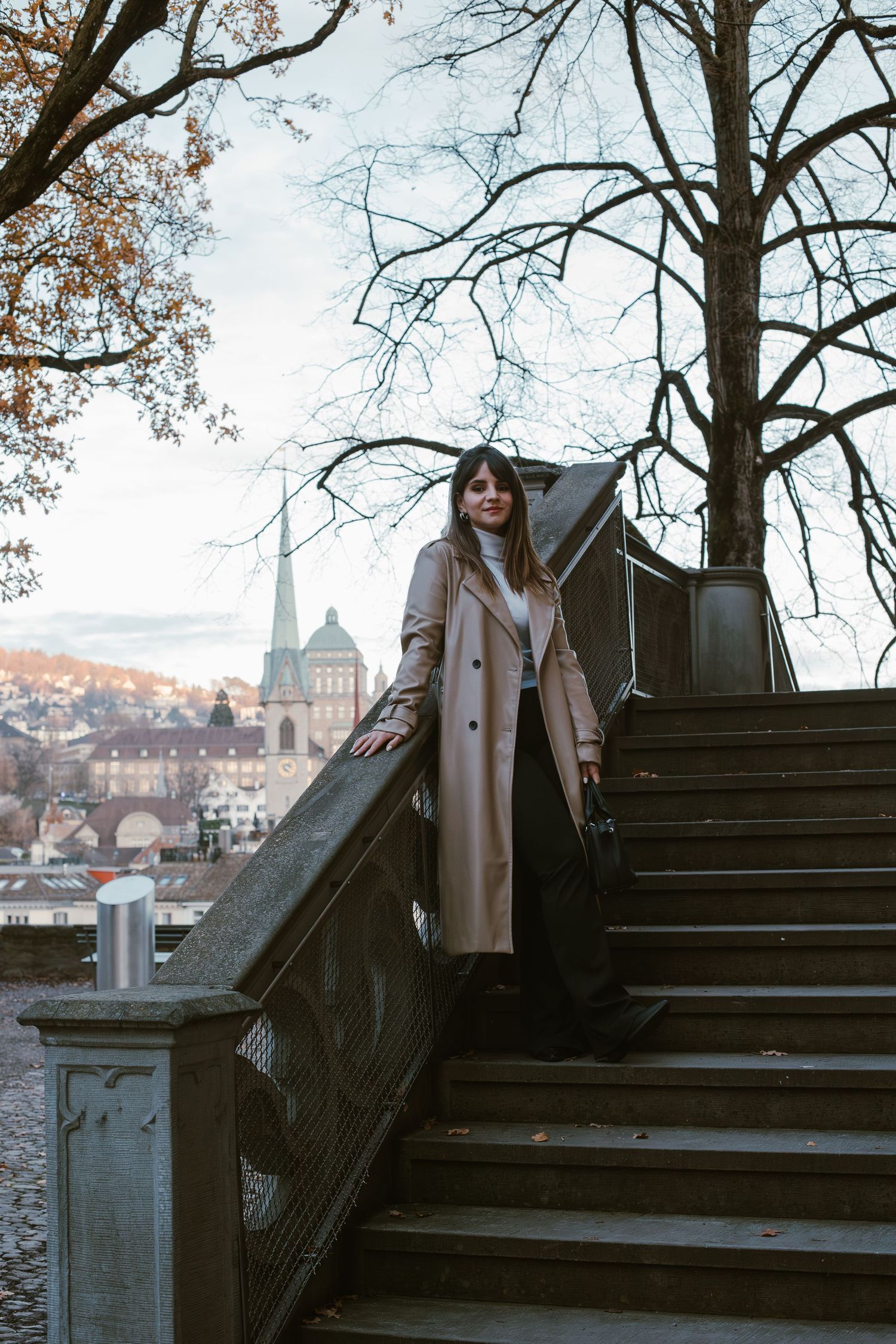 Woman in beige coat poses on stone steps with historic architecture in background.
