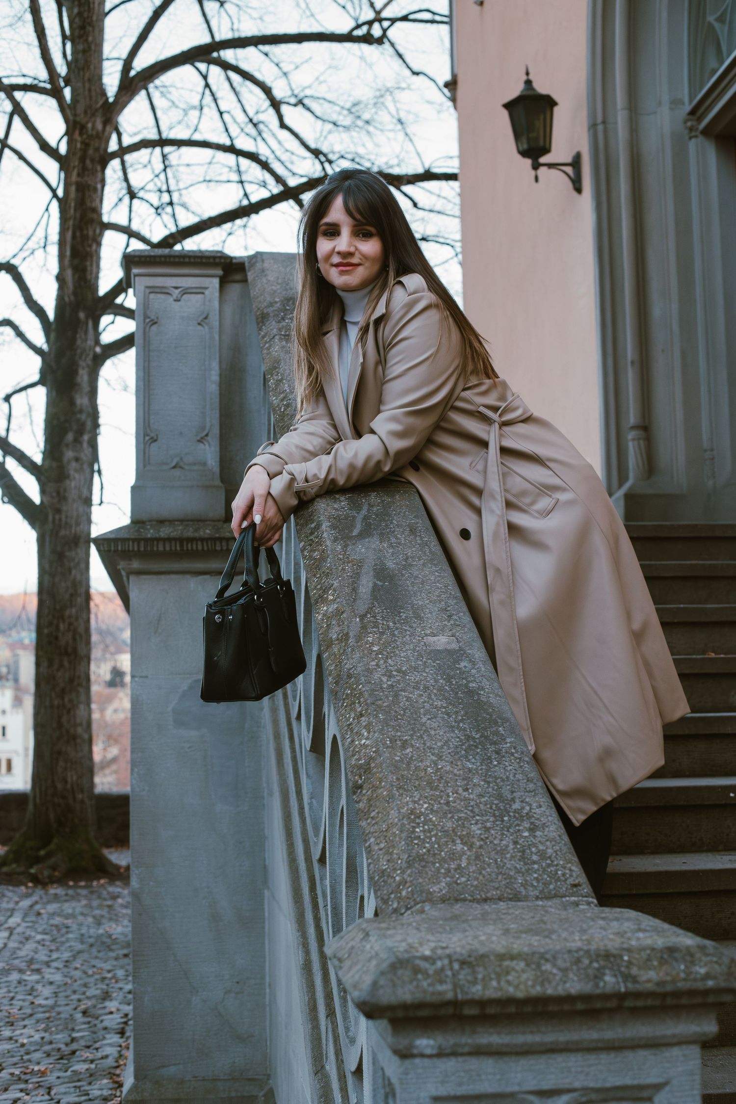 A person in a beige trench coat poses on stone steps with a black handbag against an architectural backdrop.