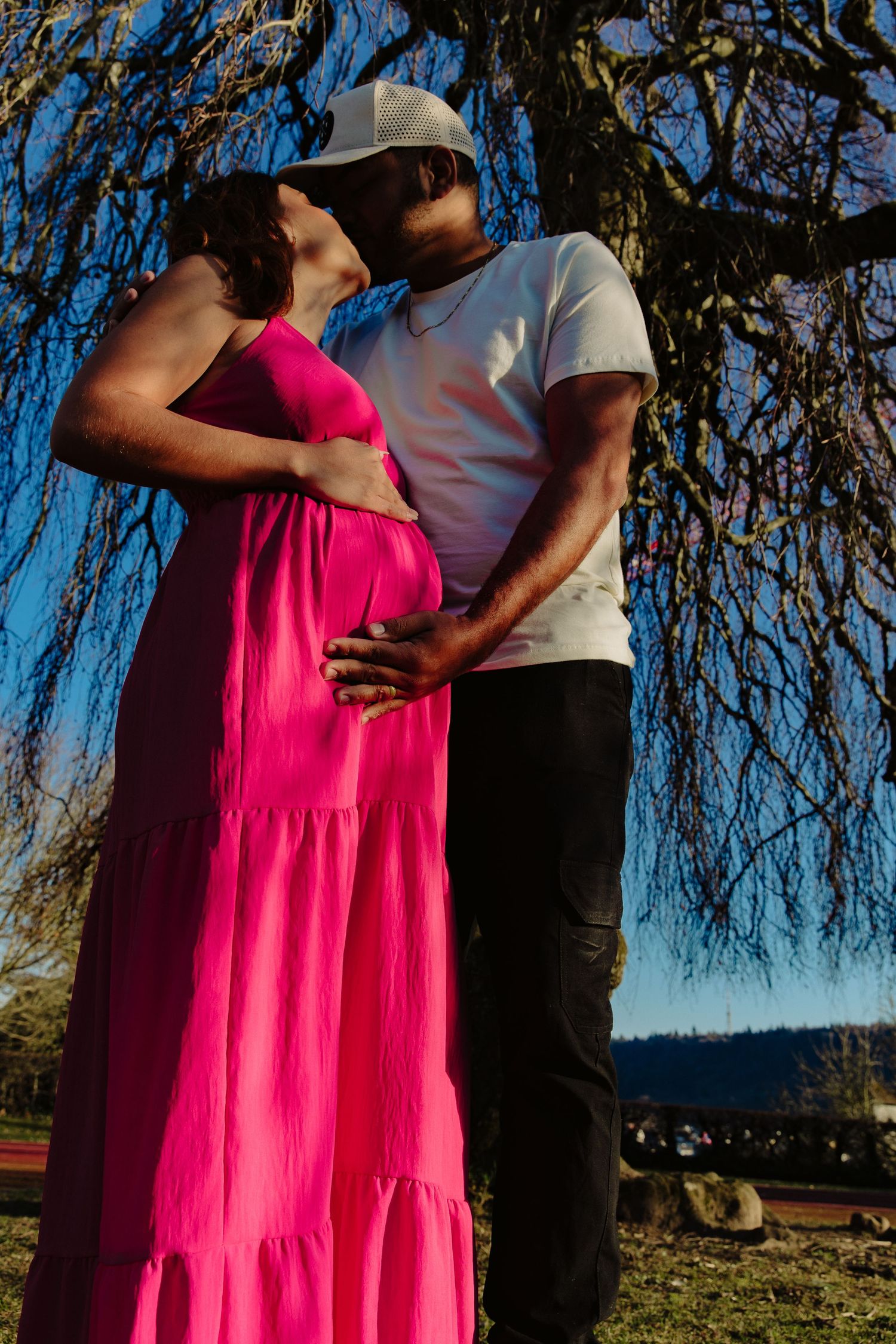 Couple embracing during pregnancy photoshoot, with woman in vibrant pink dress against willow tree.