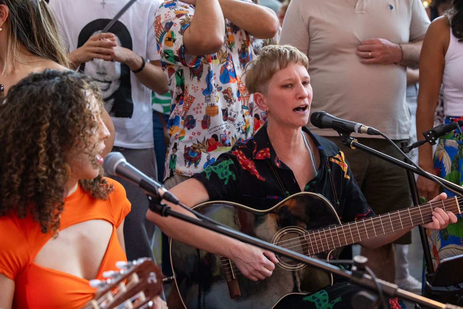 Musicians performing outdoors with acoustic guitars and microphones in front of crowd.