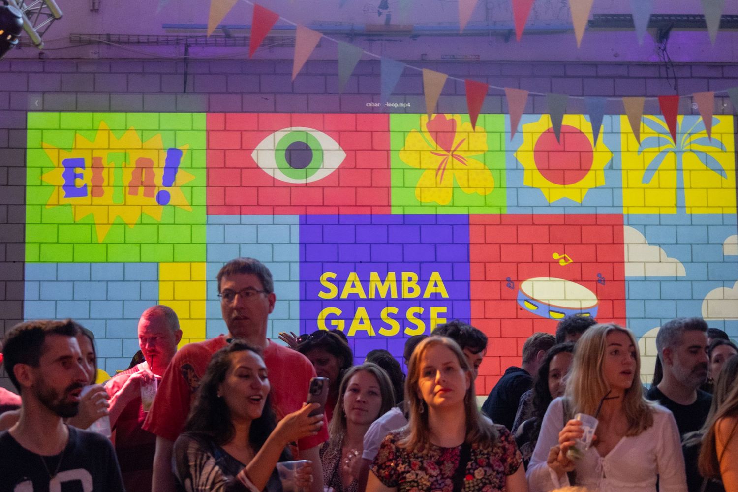 Crowd gathers under colorful wall art and bunting at Samba Gasse celebration.