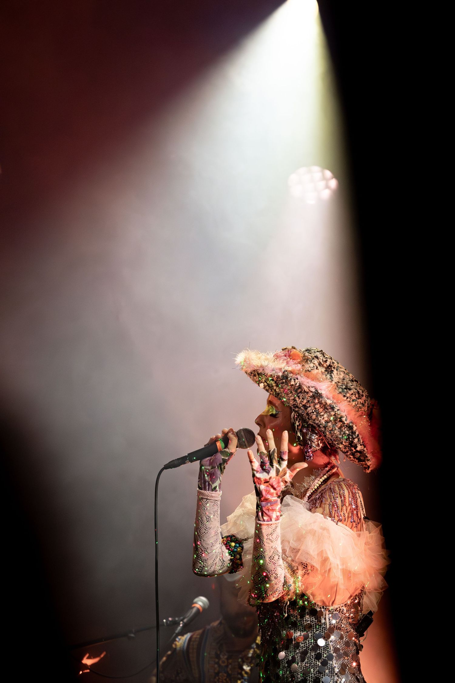 A performer in a sparkling outfit sings into a microphone under dramatic spotlight beams on stage.