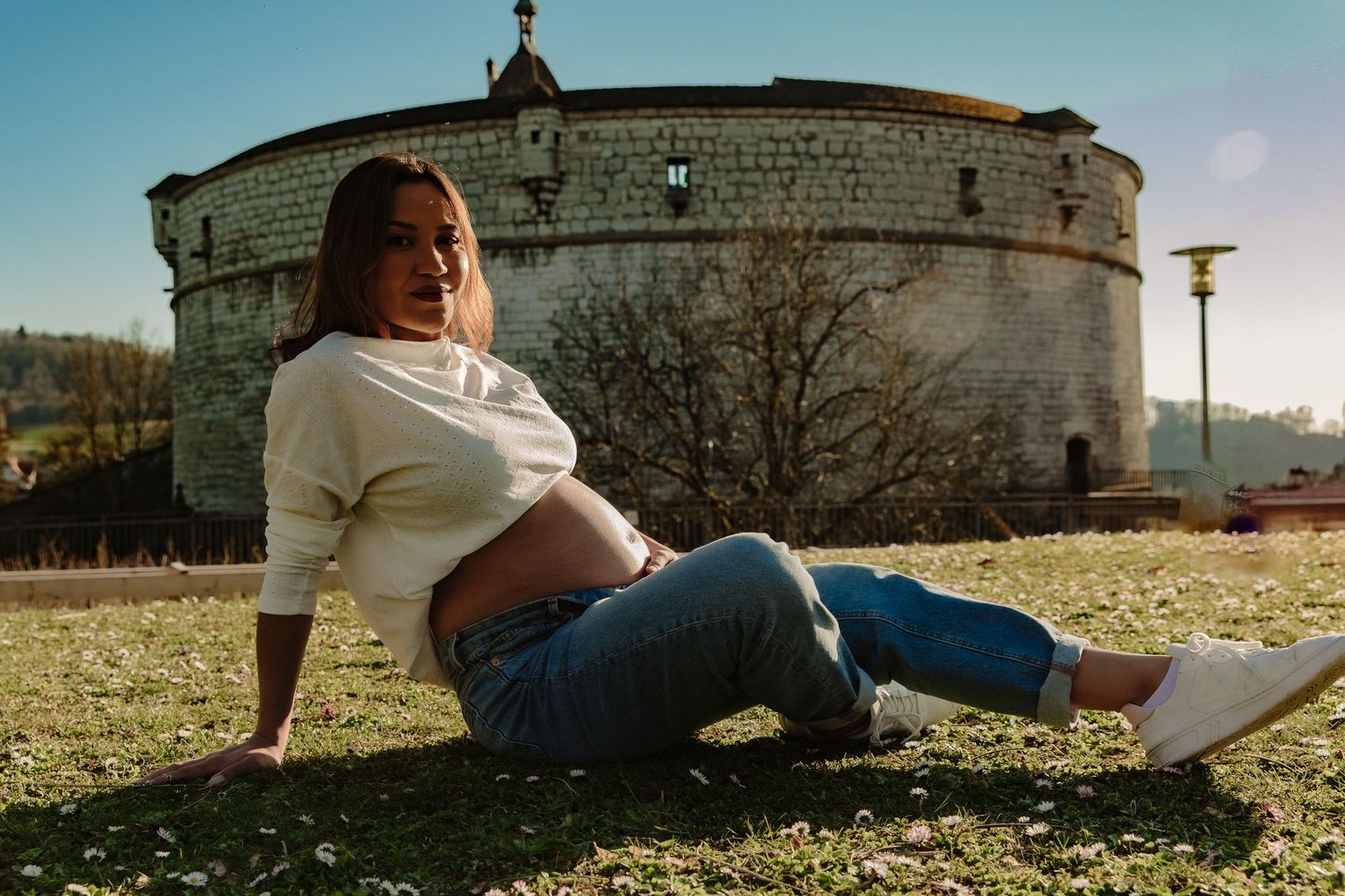 Pregnant woman relaxes on grass near historic stone fortress while wearing casual attire.