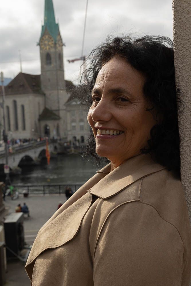 Person in beige coat smiling while overlooking historic church and river in European city.