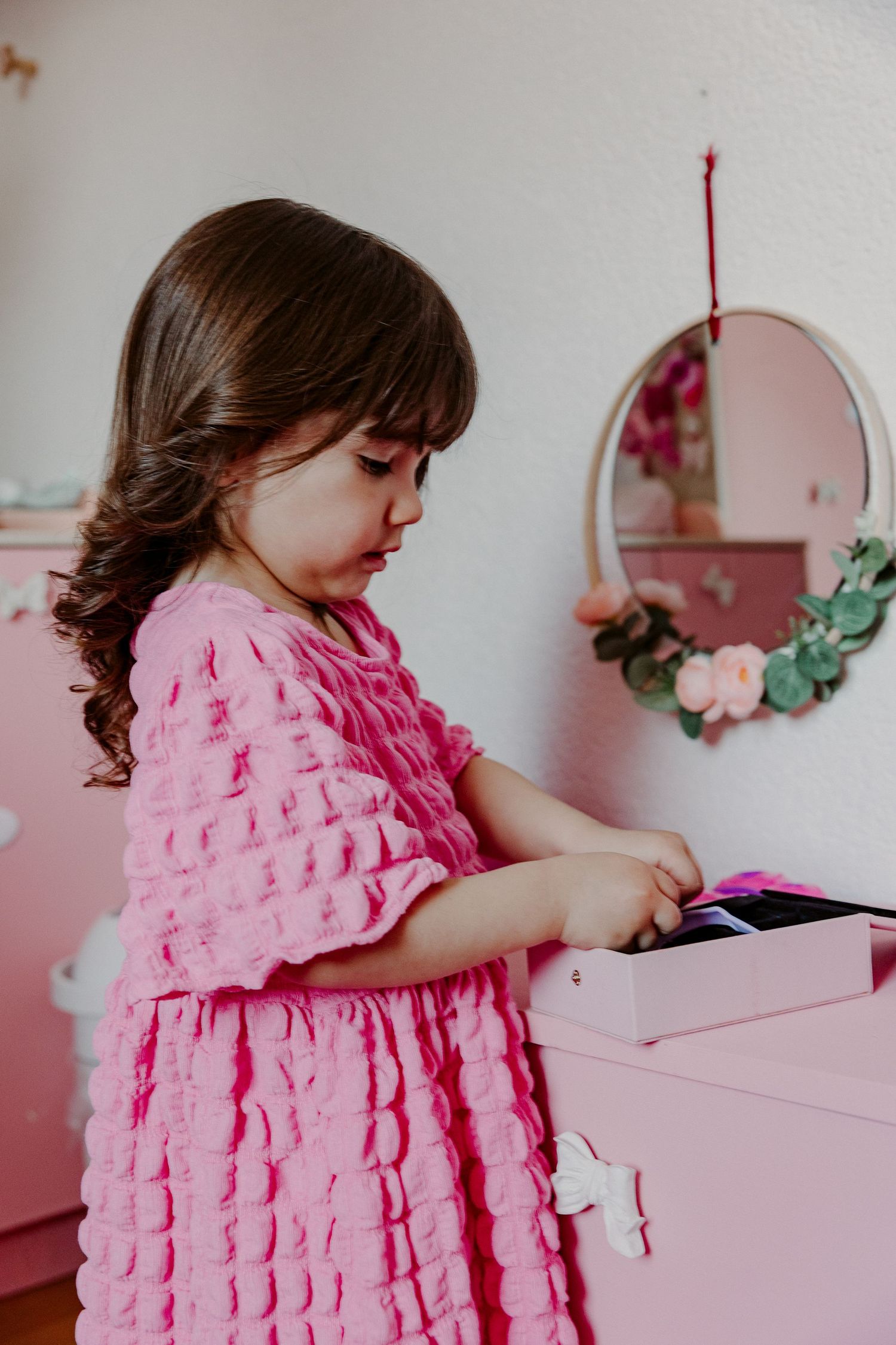 A young girl in a pink ruffled dress stands in a pink-themed bedroom.