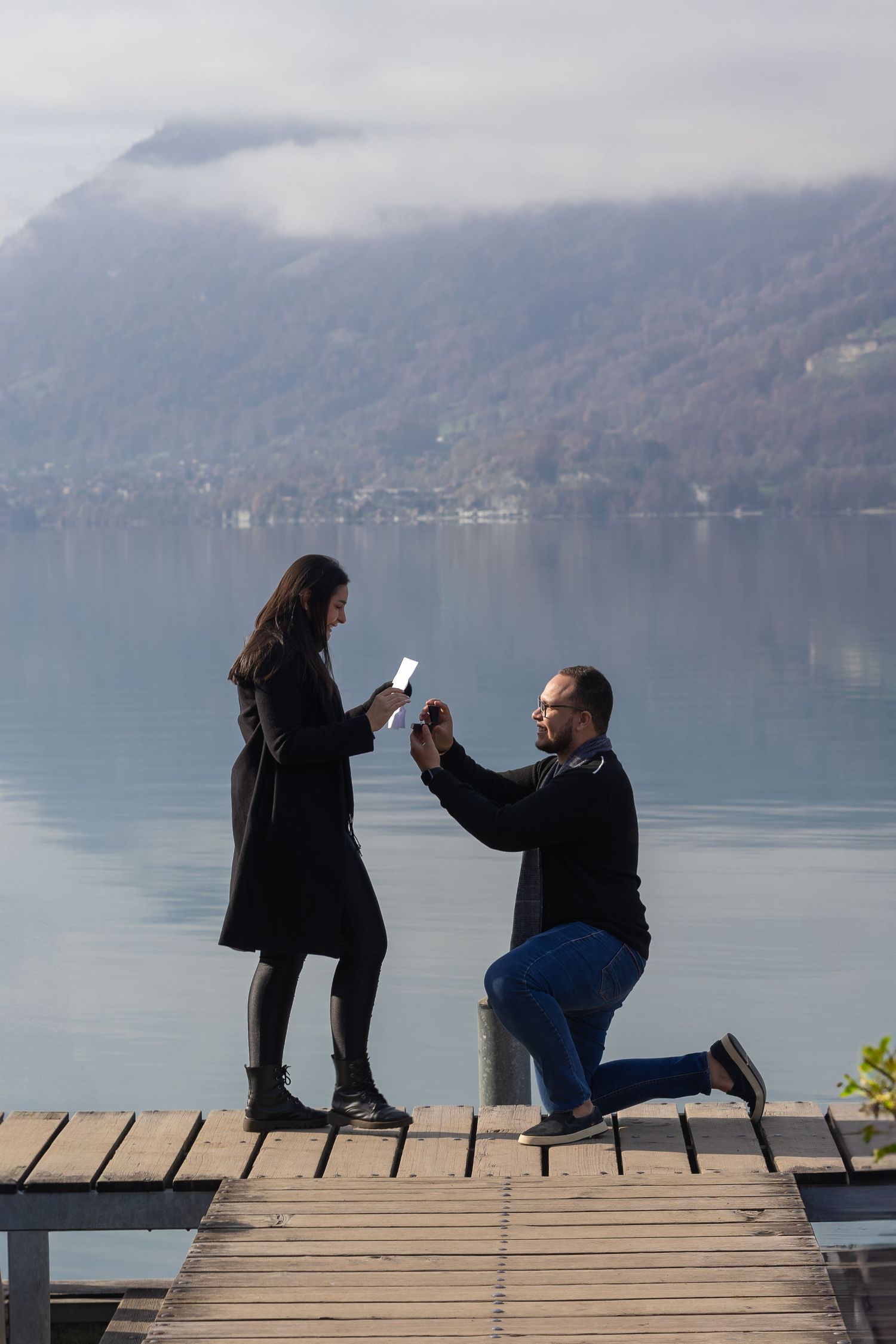 A romantic proposal takes place on a wooden pier with mountains and lake creating a stunning backdrop.