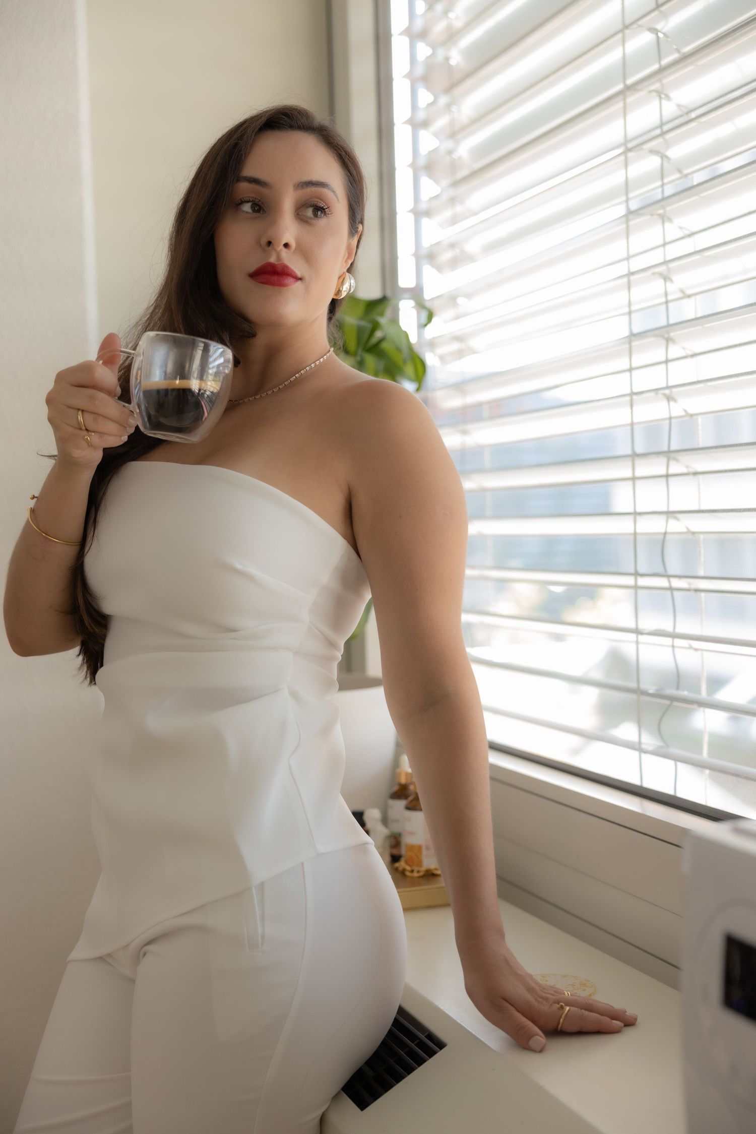 Woman in white outfit posing with coffee cup by bright window.