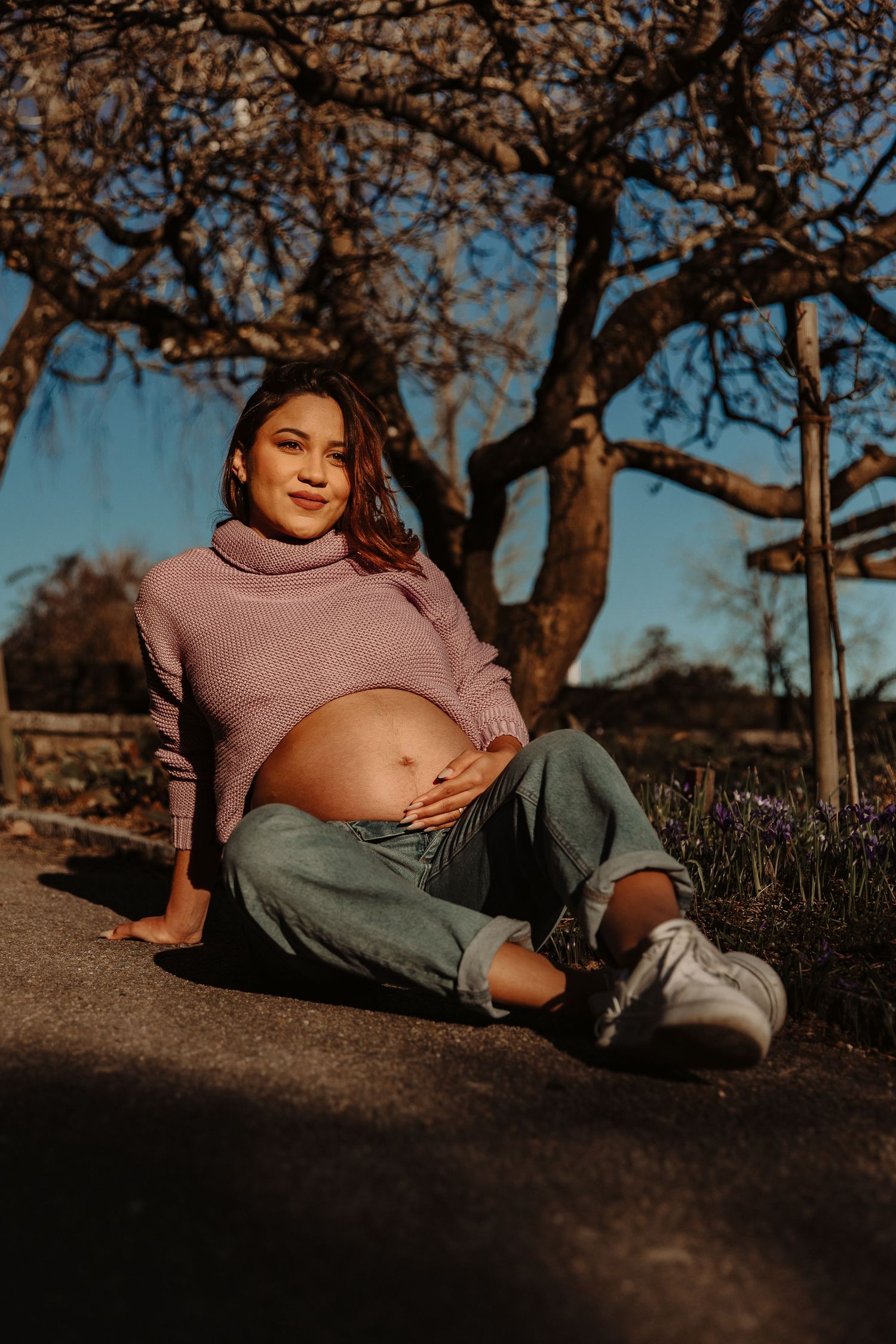 Pregnancy photo session outdoors with woman in mauve turtleneck sweater sitting under tree.