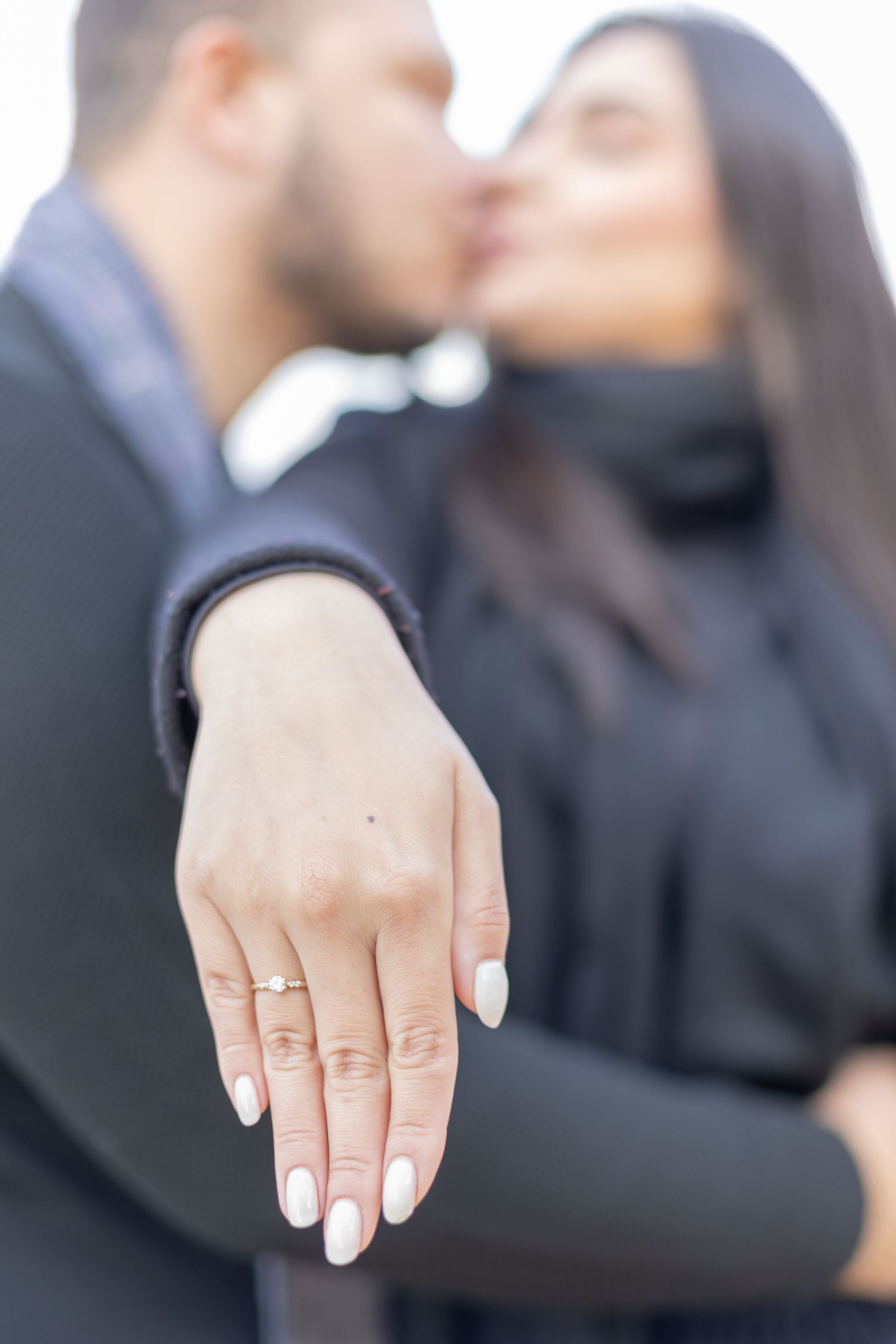 A close-up of a hand displaying an engagement ring while a couple kisses in the background.