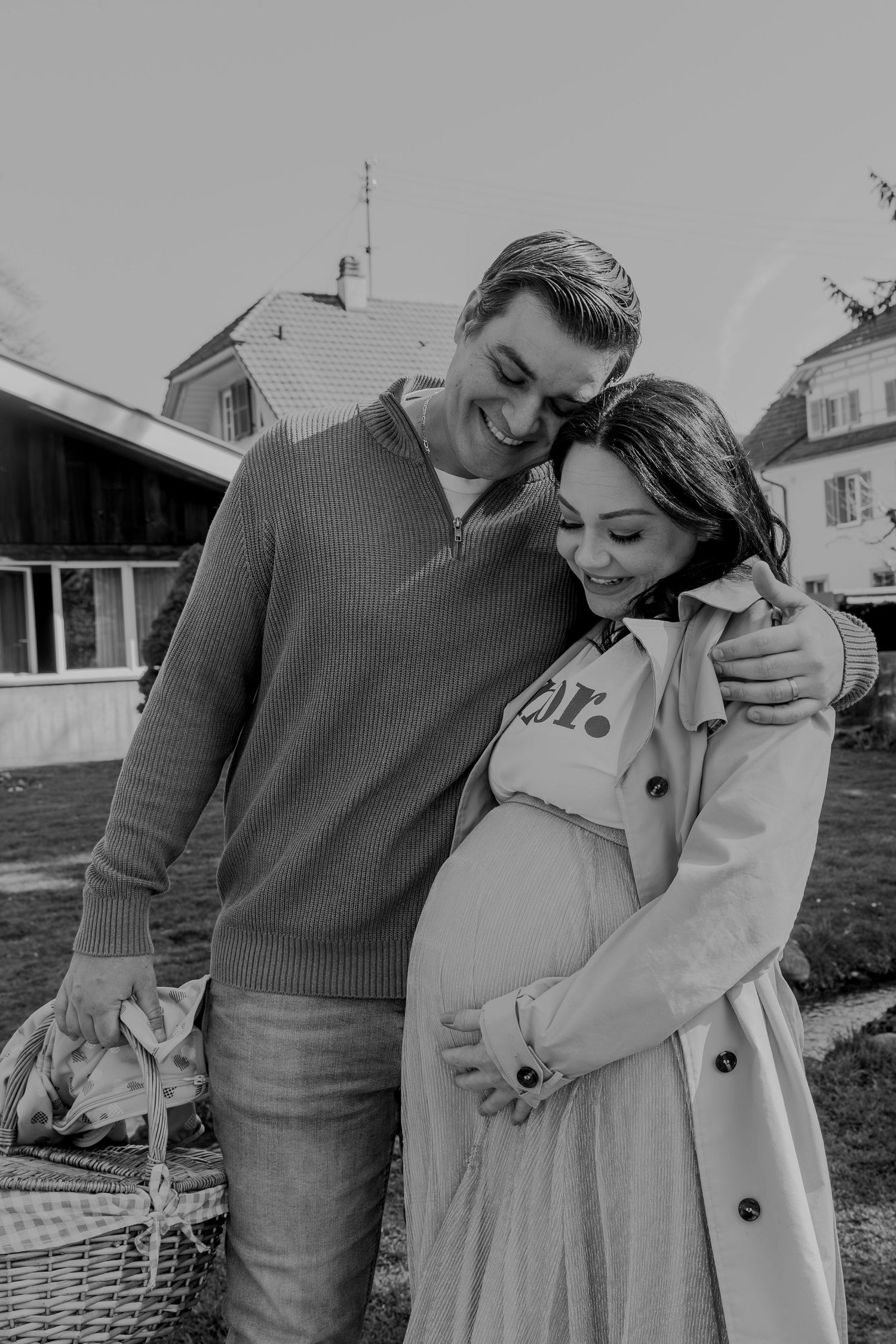 A black and white portrait of an expecting couple sharing an intimate moment while holding a picnic basket.