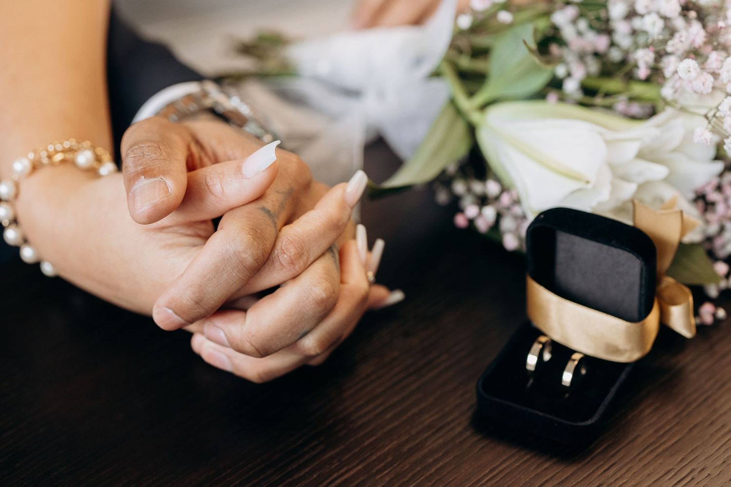 Two hands clasped together next to an engagement ring box on a dark wooden surface with flowers in the background.