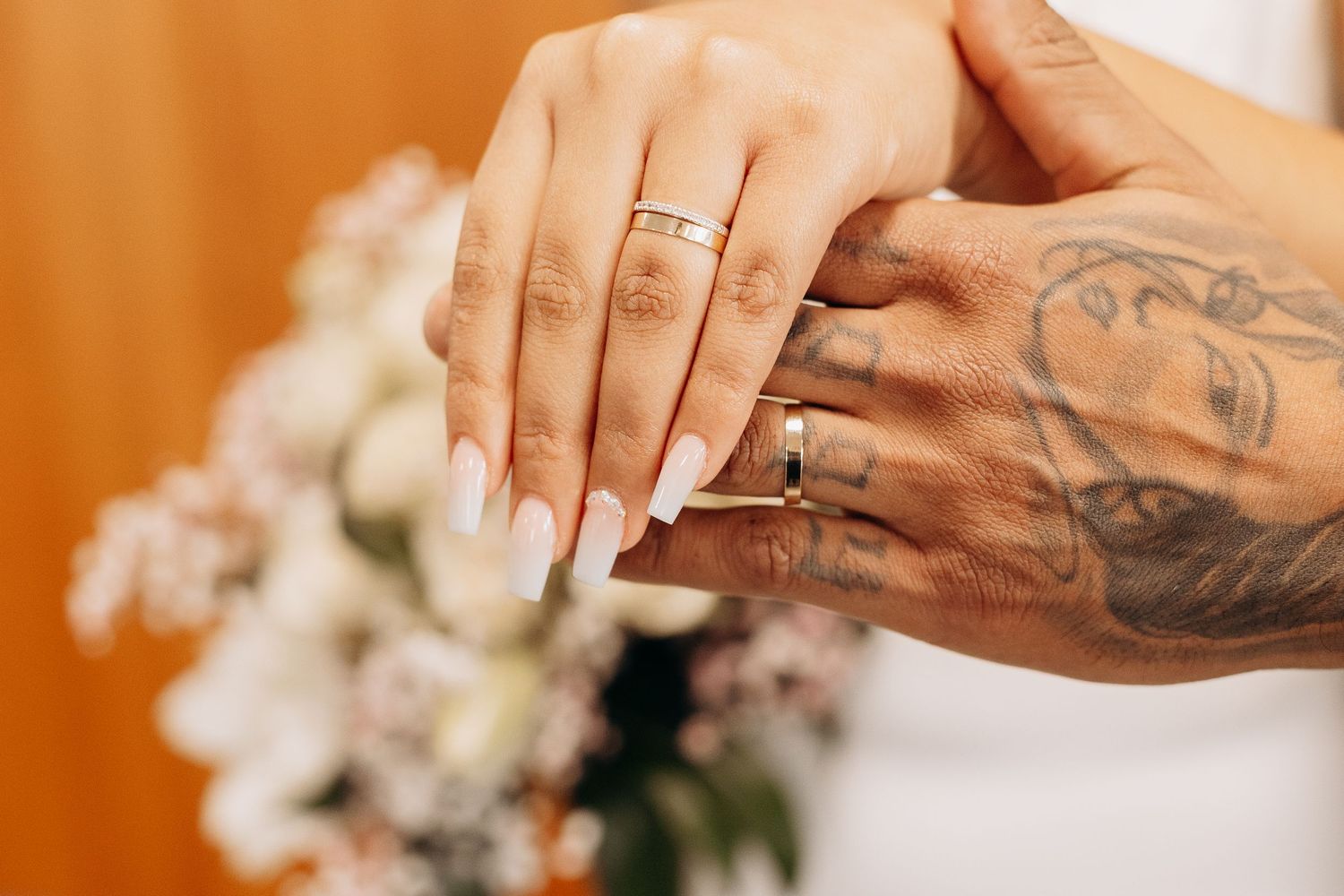 Close-up of hands exchanging wedding rings with tattoos and floral bouquet in background.