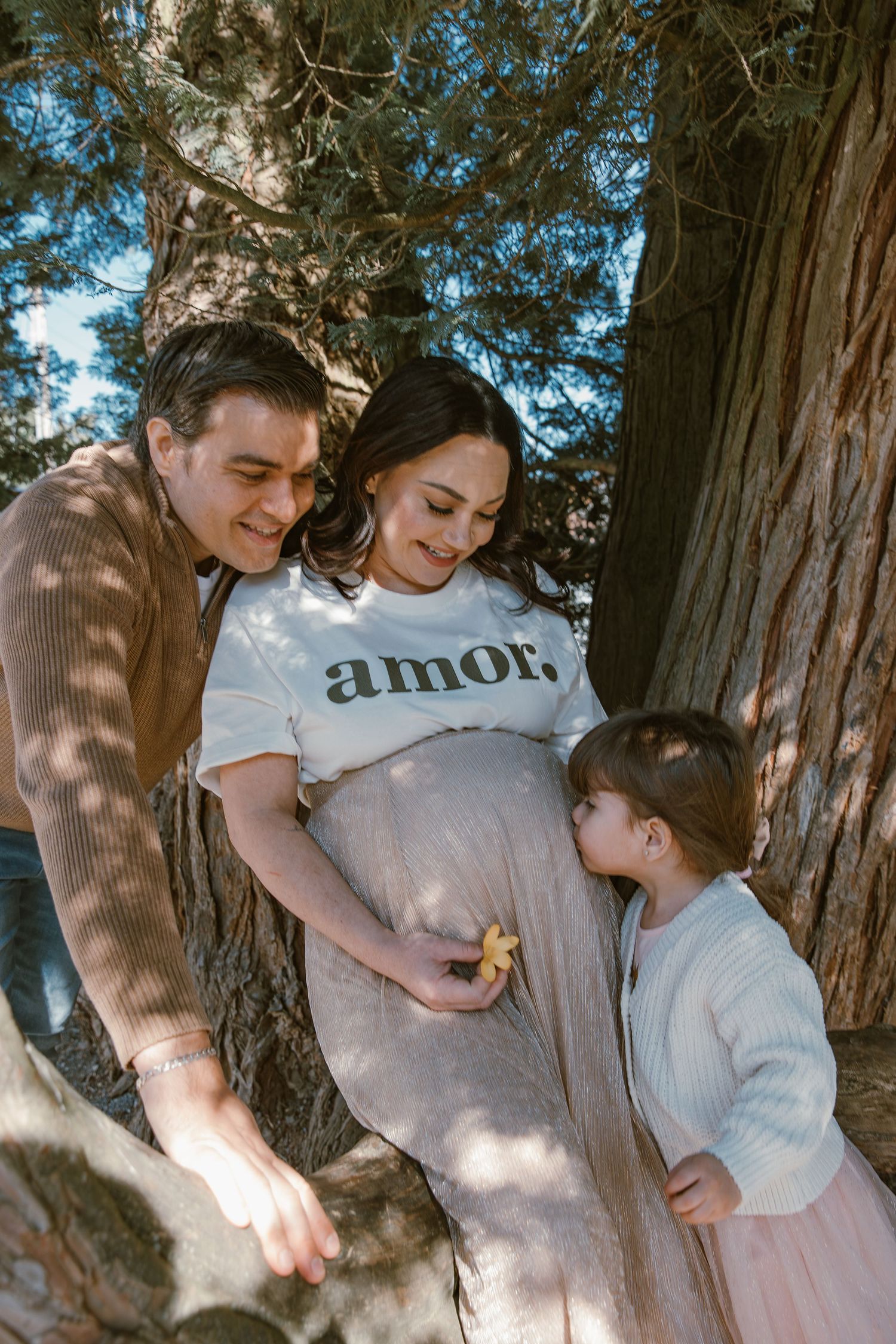 Family maternity photo under trees with pregnant mother wearing amor shirt and child touching belly.