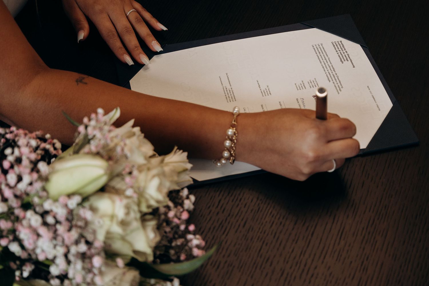 Hands signing marriage documents next to delicate floral arrangements on wooden table.