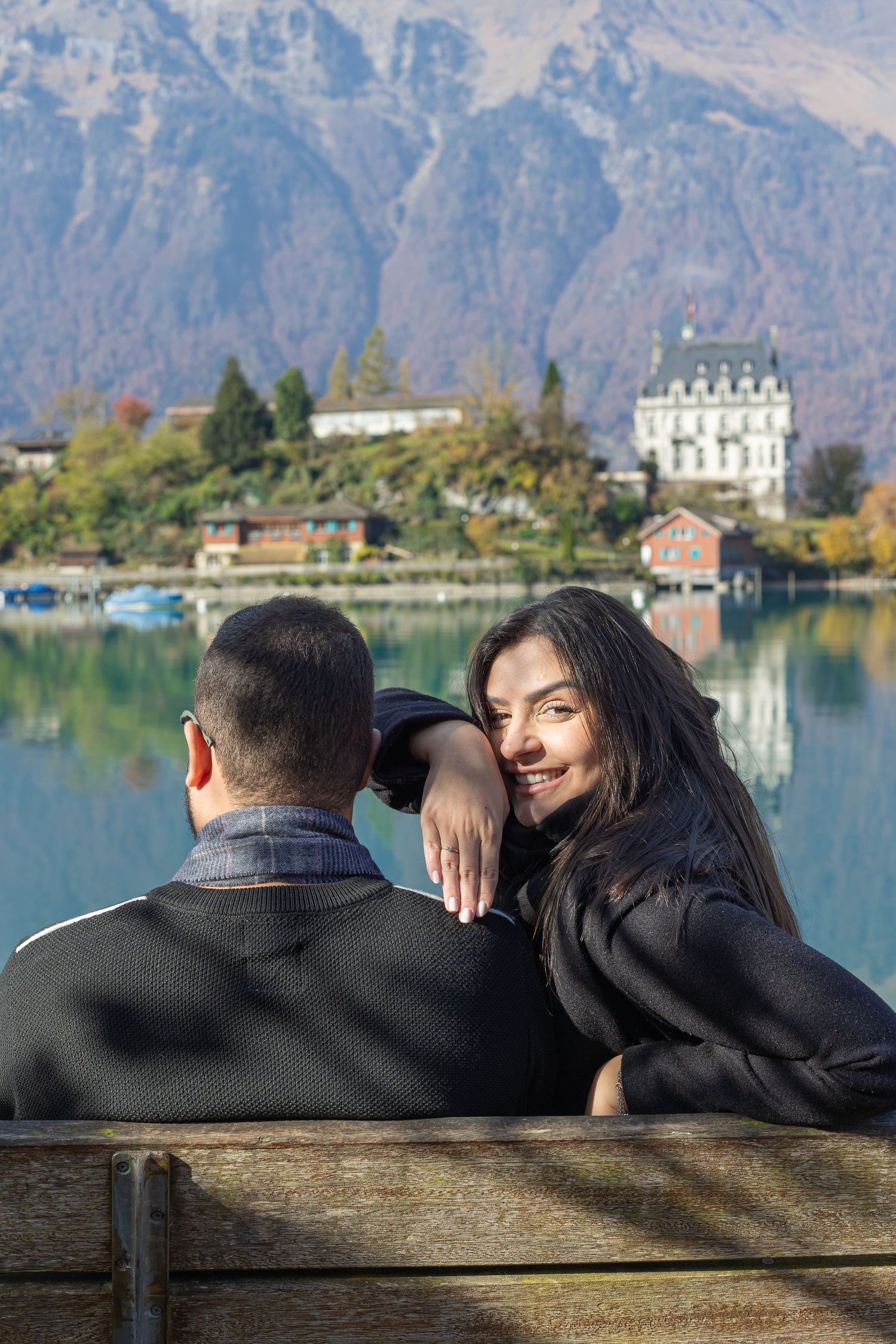 A couple enjoys a romantic moment on a bench with a scenic lakeside chateau in the background.