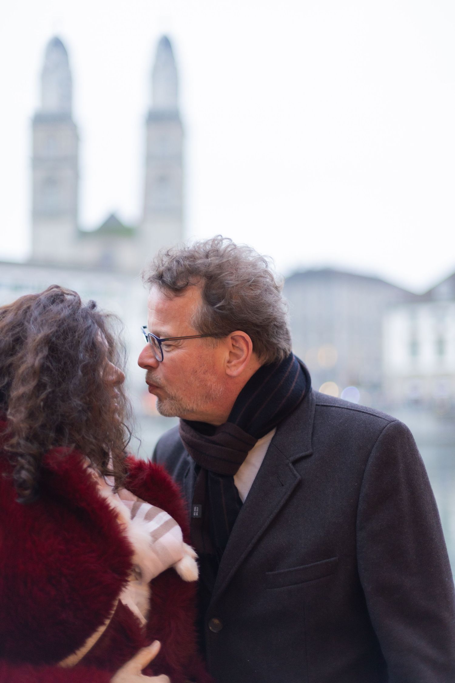 Two people in formal winter attire share a close conversation with city architecture in background.