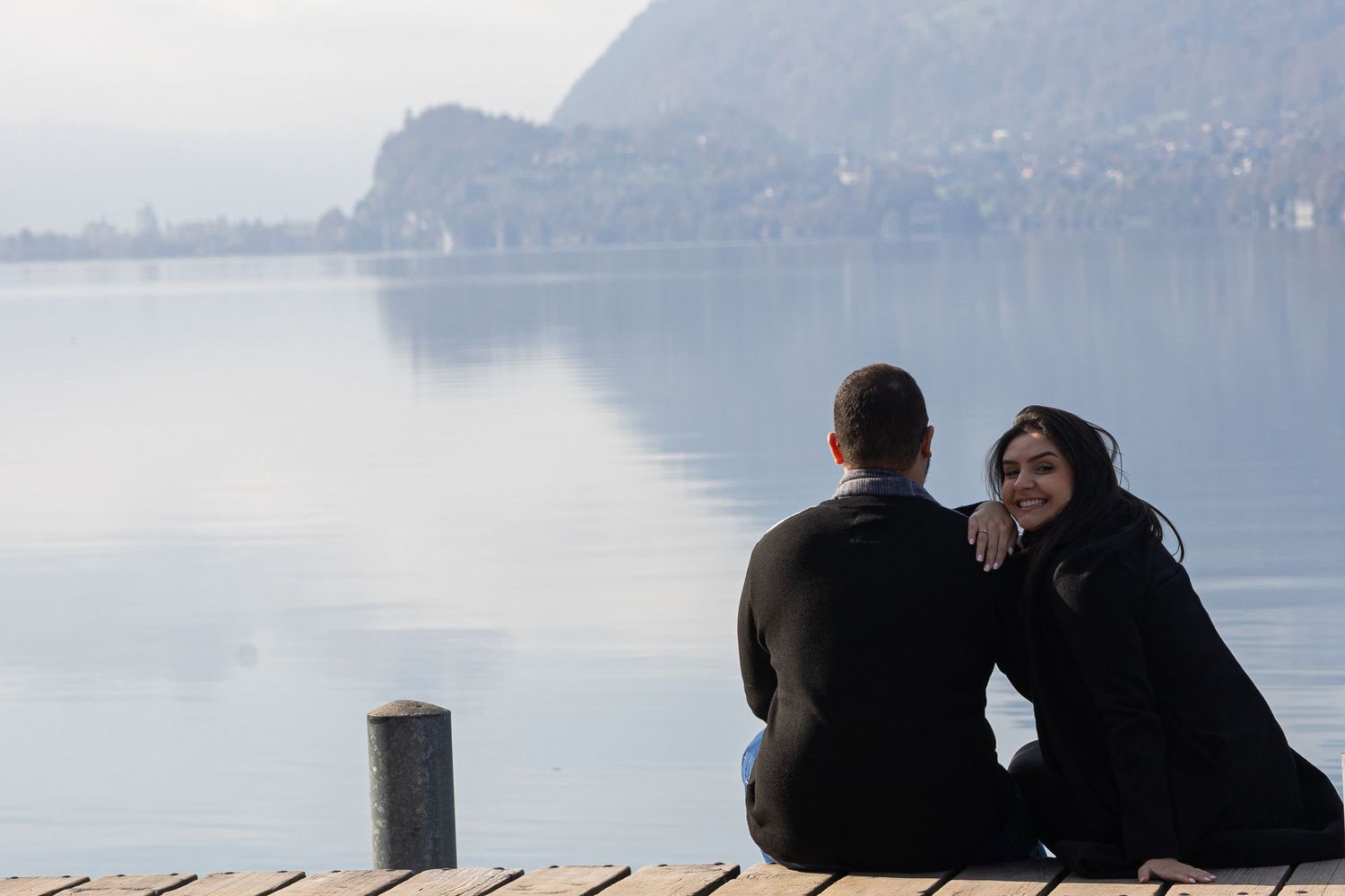 A couple sits together on a wooden dock overlooking a serene lake with mountains in the background.